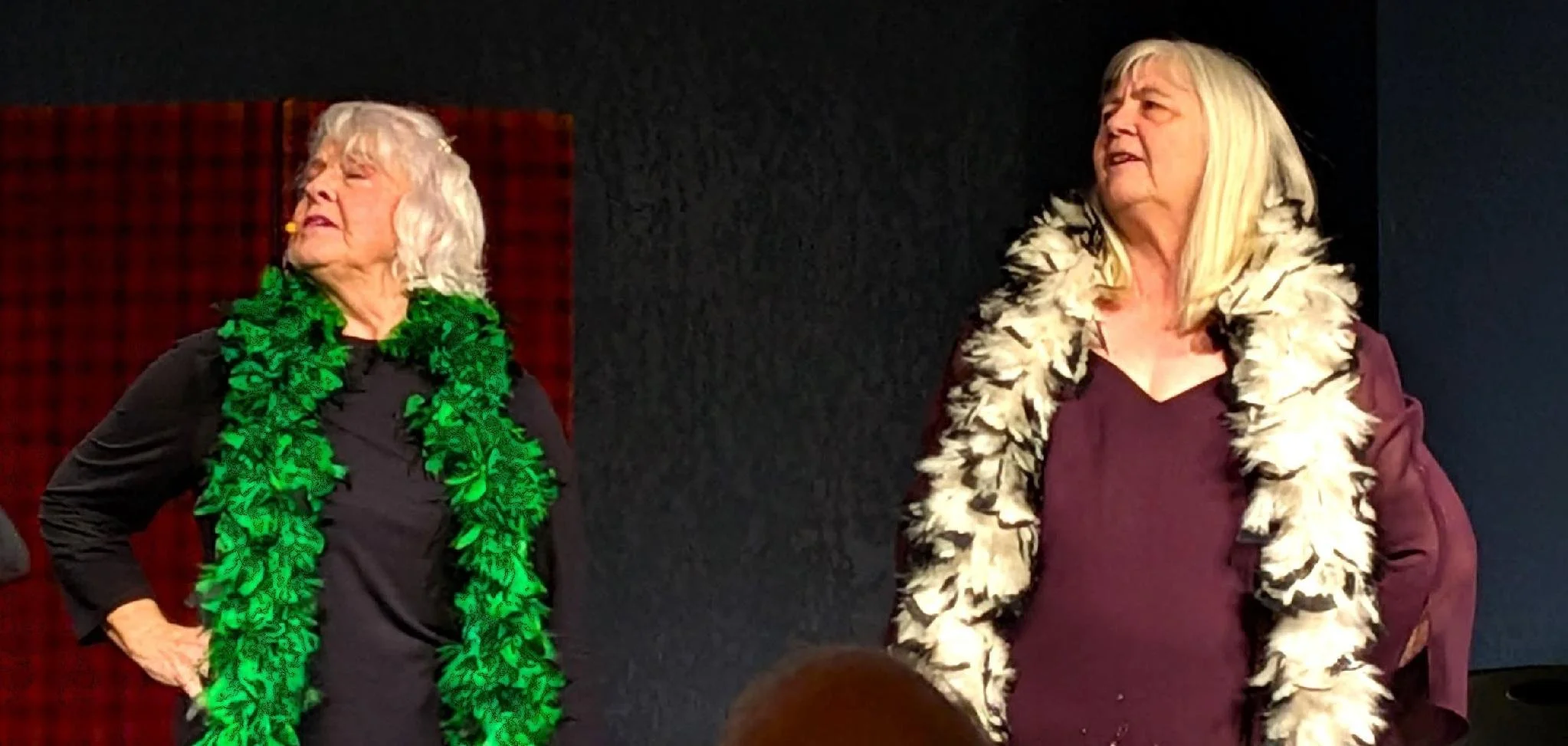 Two elderly women standing on stage, one wearing a green leafy scarf and the other a cream feathered scarf, both looking to the side.