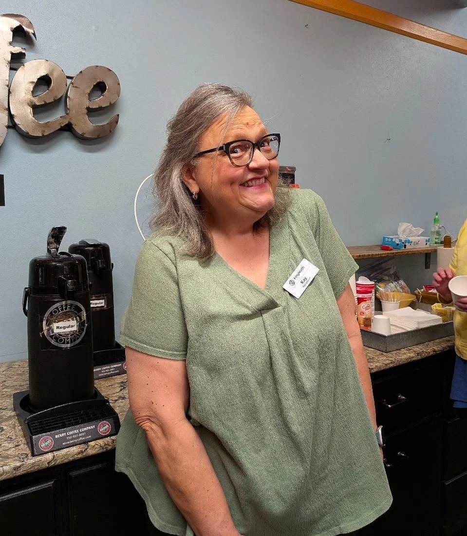 A woman with gray hair, black glasses, and a green top, smiling in a coffee shop setting near a counter with coffee dispensers, cups, and snacks.