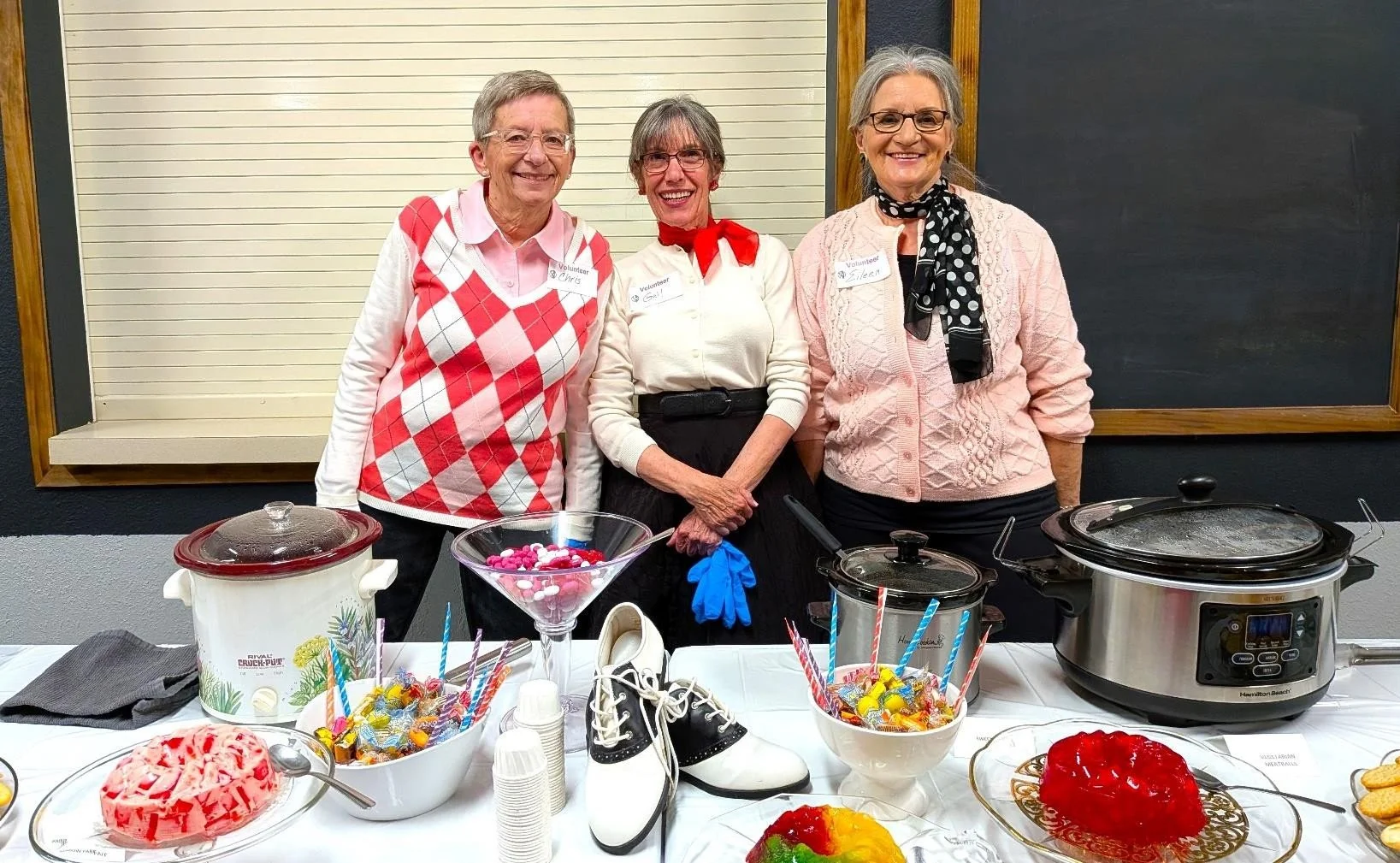 Three women standing behind a table with desserts and crockpot, smiling at the camera. They are inside a room with black and wood-paneled walls. The women are wearing name tags and vintage clothing accents, with one holding a pair of blue gloves. The table has various candies, a pair of ice skates, and slices of cake or pie.