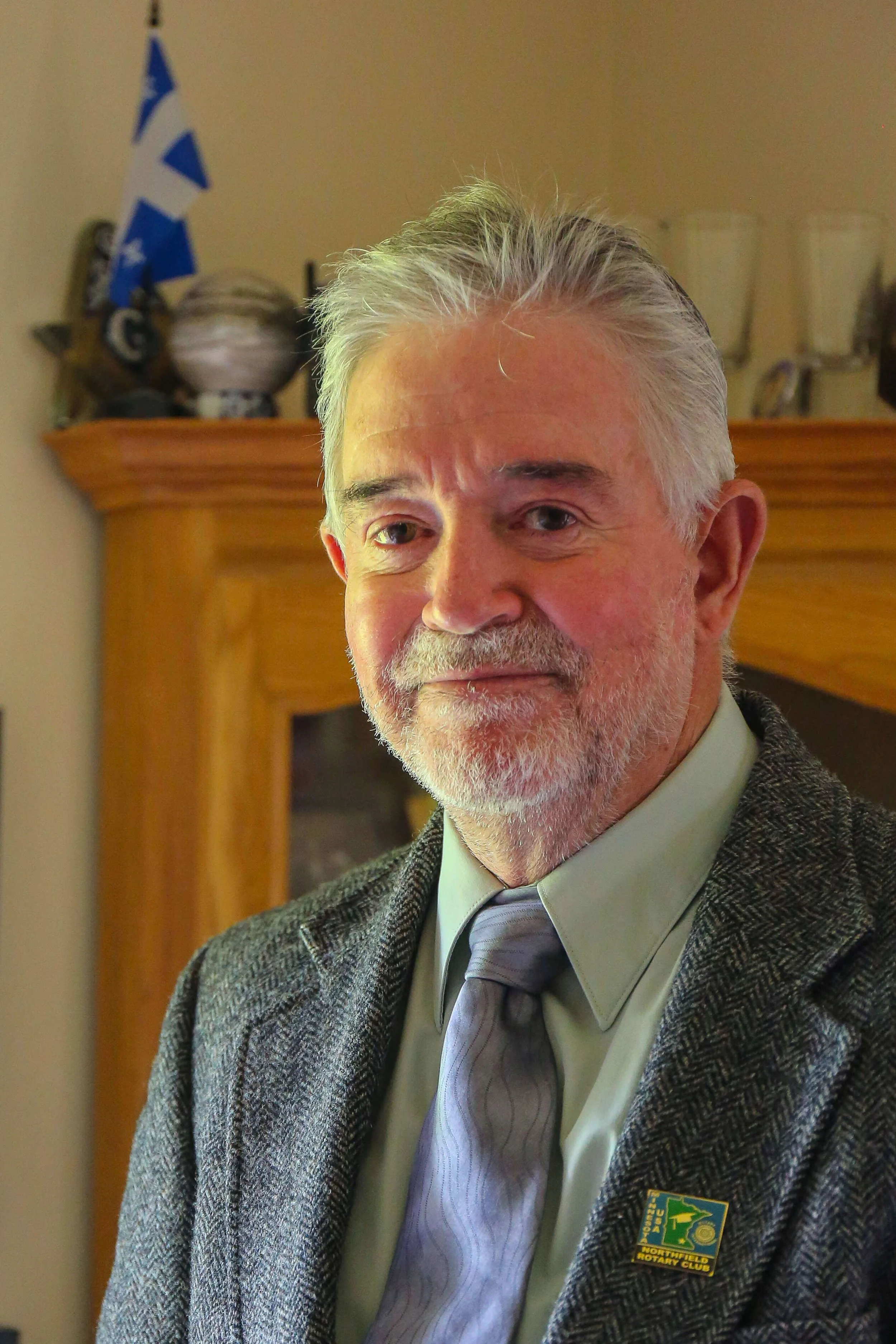A man in a gray tweed blazer, light green shirt, and purple tie stands in front of a wooden cabinet with trophies and a small blue and white flag. The blazer has a badge that reads 4-H Northfield Rotary Club.