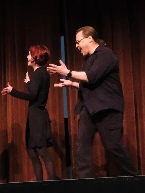 Two people, a woman with short red hair and a man with glasses, are performing on stage with dark curtains behind them. They are smiling and gesturing with their hands.