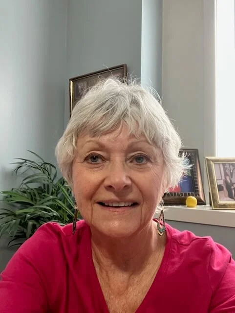 A smiling elderly woman with short gray hair, wearing earrings and a pink top, indoors with framed photos and a plant in the background.