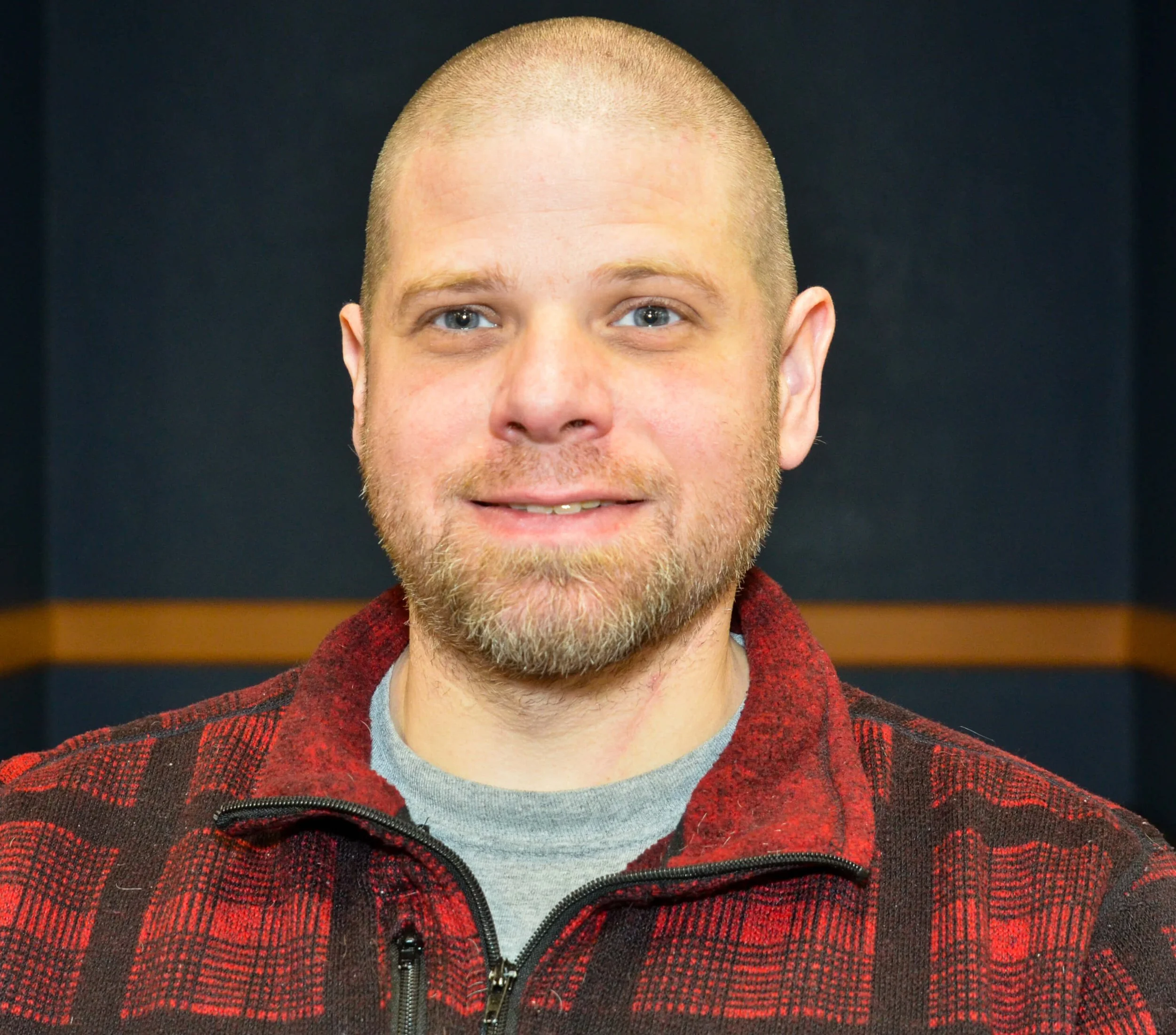 A close-up portrait of a man with light skin, short blond hair, and blue eyes, smiling slightly, wearing a red and black checkered jacket over a light gray shirt, against a dark background.