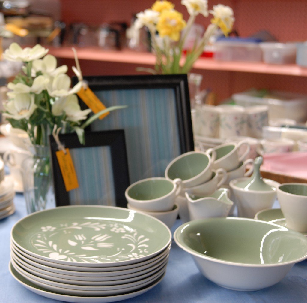 Display of green and white ceramic dinnerware, including plates, bowls, and cups, arranged on a table with floral arrangements and picture frames in the background.