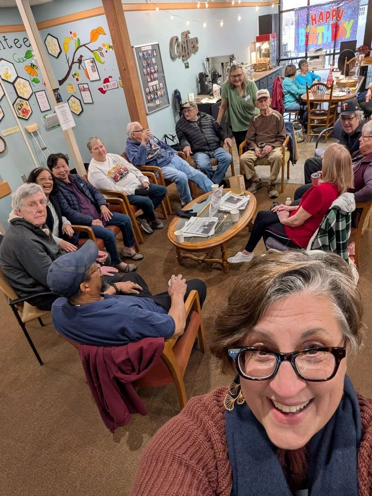 Group of people gathered in a cozy room for a birthday celebration, with a large 'Happy Birthday' banner hanging on the window, and various wall decorations, including framed photos and a colorful tree mural. Some people are seated around a coffee table with newspapers and cups, smiling and engaging with each other.