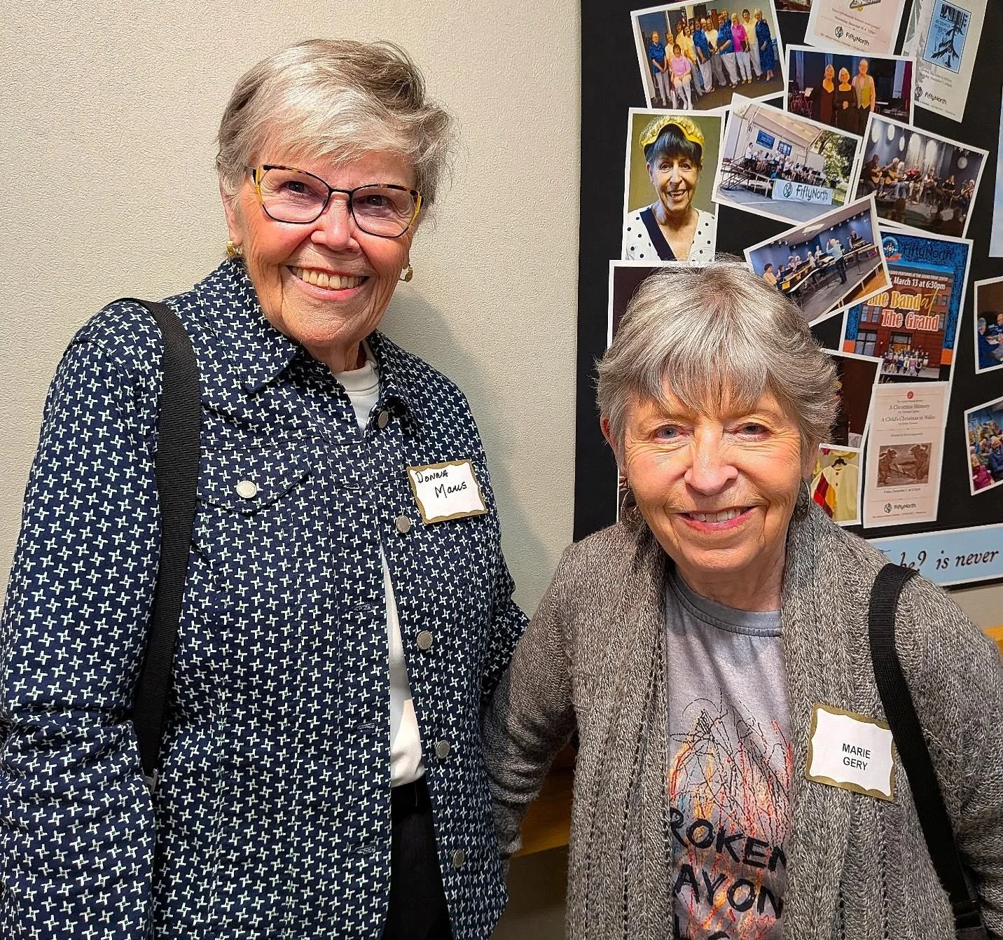 Two elderly women smiling for the camera at an indoor event, standing in front of a black poster with photos and various images pinned to it. The woman on the left wears glasses, a patterned navy and white jacket, and has a name tag reading 'Donna Maus'. The woman on the right wears a gray jacket over a T-shirt with a graphic design, and has a name tag reading 'Marie Gery'.