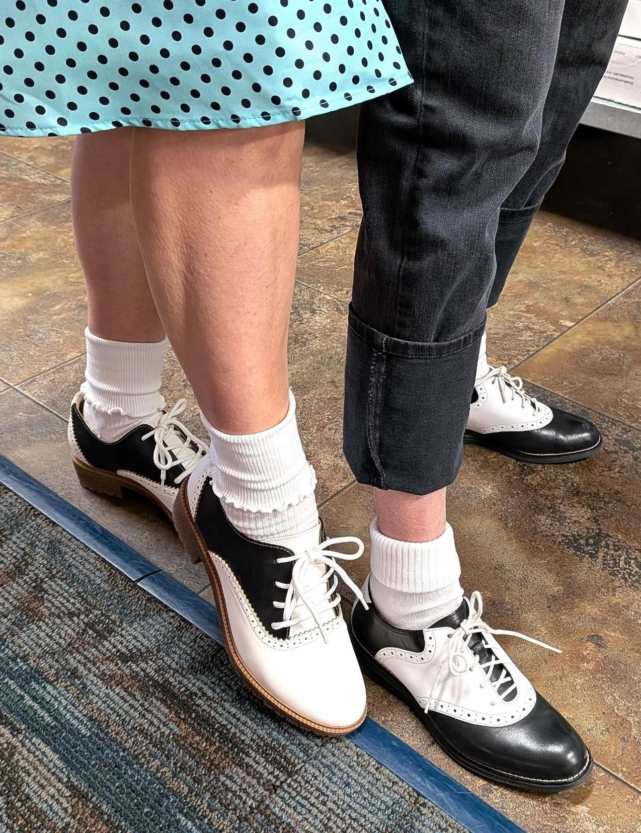 Close-up of two people with their legs touching; one person wearing black and white two-tone shoes with white socks, and the other wearing black jeans and black-and-white shoes, standing on a tiled floor.