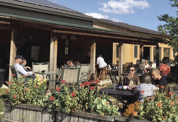 People dining outside at a patio with flowering plants in the foreground, and a wooden building with a porch in the background.