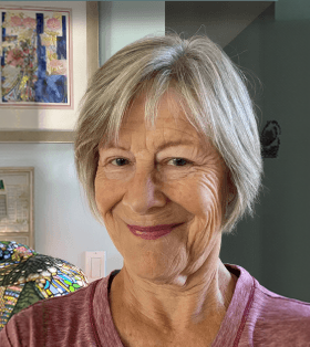 A smiling older woman with short gray hair wearing a pink top, standing indoors in front of a wall with artwork and a colorful quilt visible.