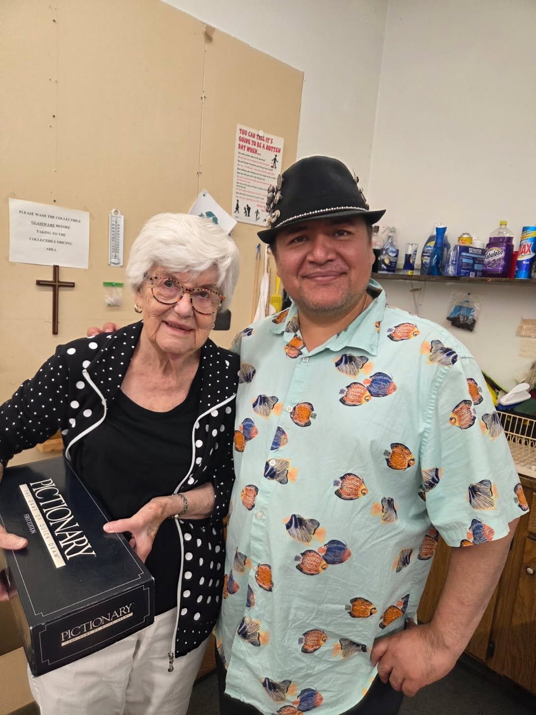 An elderly woman with white hair, glasses, and a black polka-dot jacket, standing next to a man wearing a black hat with embellishments and a colorful fish-patterned shirt. The woman is holding a black box labeled "PICTIONARY". They are indoors, with shelves and signs in the background.
