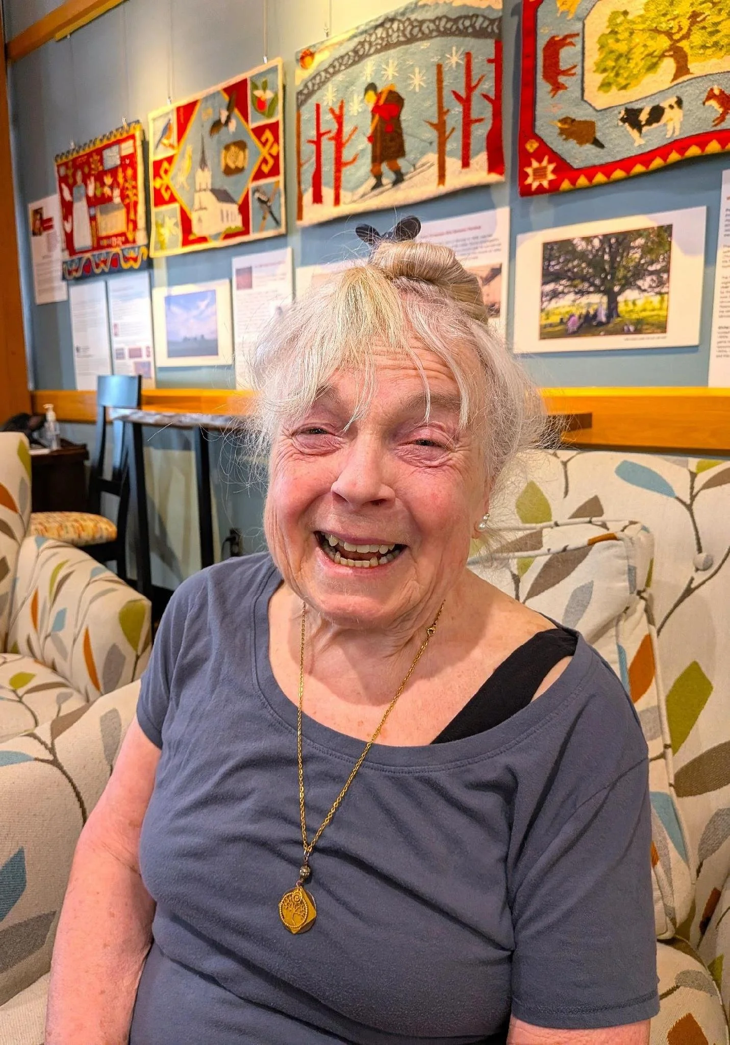 Smiling elderly woman with gray hair tied up in a bun, wearing a dark gray shirt with a black strap visible on her shoulder, a gold necklace, and a gold pendant. She is sitting on a patterned sofa in a room with framed quilt artworks on a light blue wall behind her.