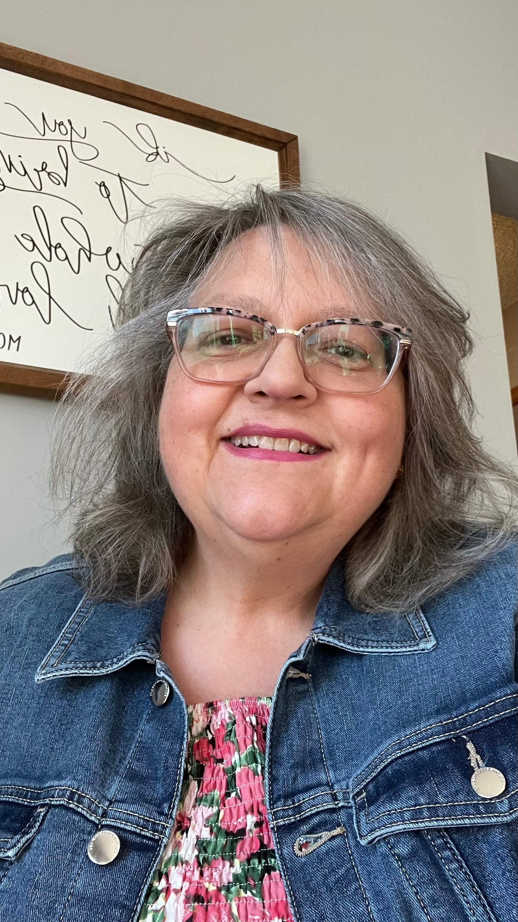 A smiling woman with gray hair, wearing glasses, a denim jacket, and a floral top, standing indoors with a whiteboard in the background.