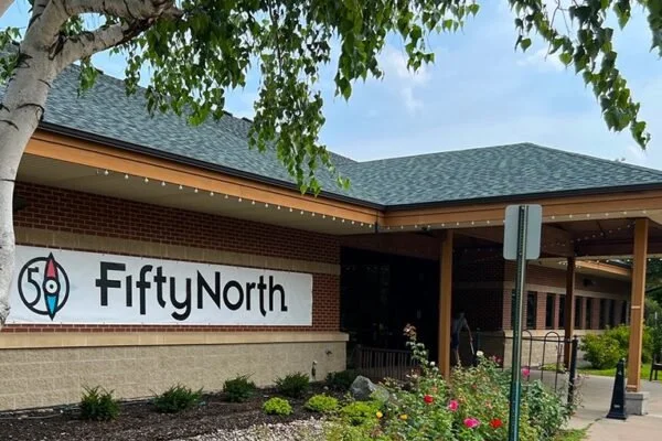 Sign for Fifty North restaurant outside a brick building with a green roof, surrounded by flowers and greenery.