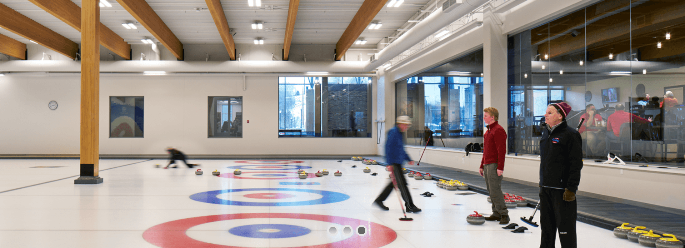 Indoor curling rink with people playing and coaches watching, curling stones on the ice, and a large glass window showing its surroundings.