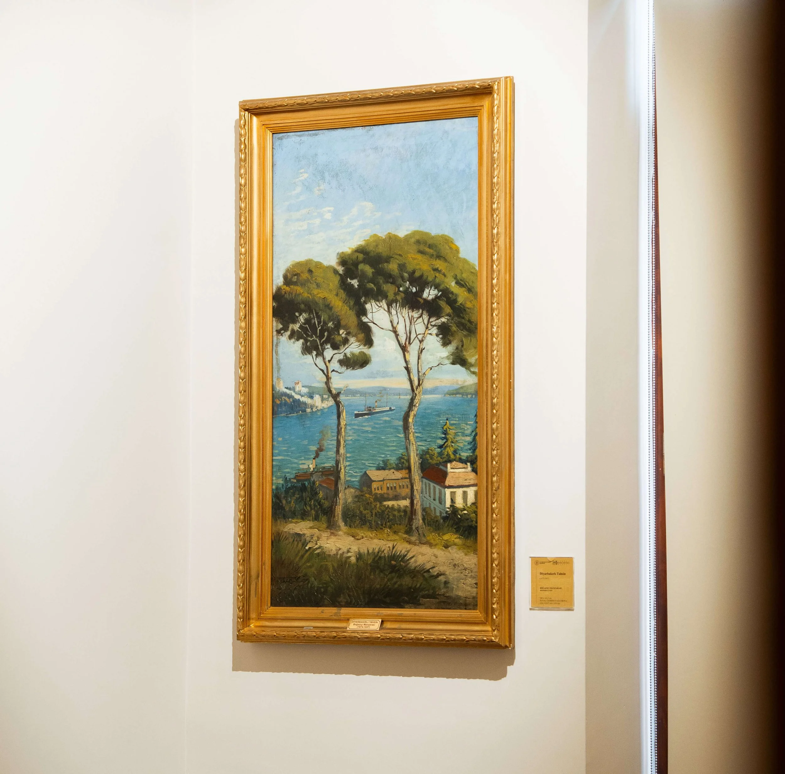 Landscape painting of a lakeside scene with trees and a boat, inside a gold ornate frame, on a white gallery wall.