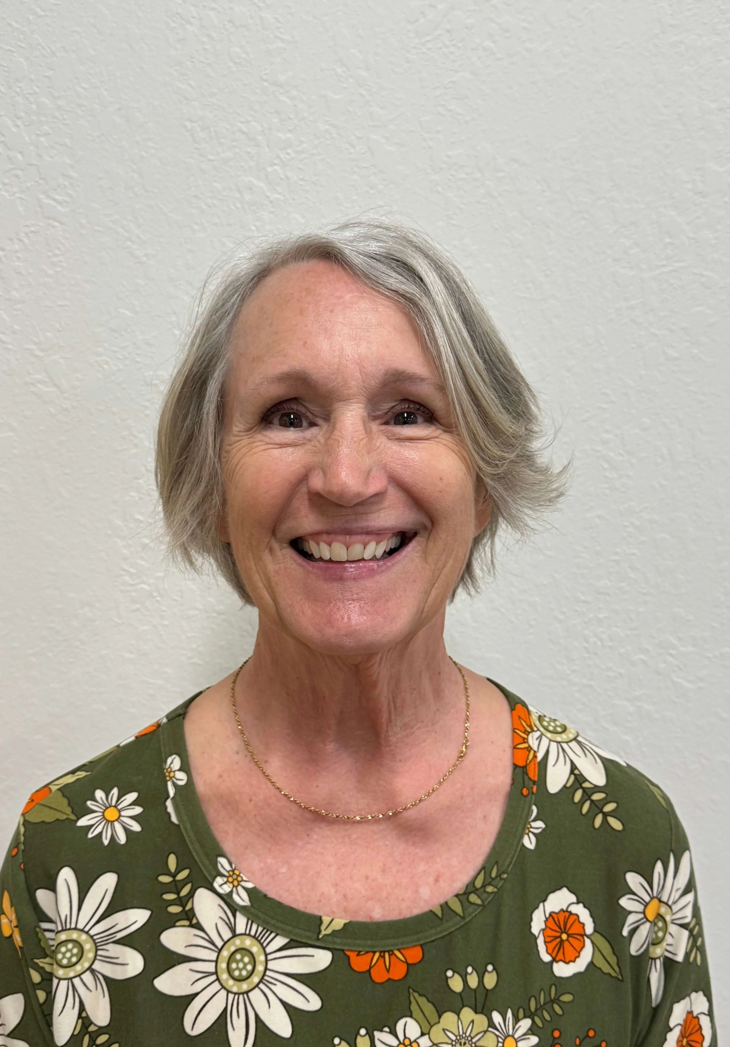 A smiling middle-aged woman with short gray hair, wearing a green floral top and a gold necklace, against a plain white wall background.