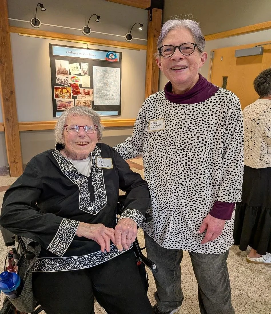 Two women smiling, one in a wheelchair wearing a black embroidered top, and another standing wearing glasses and a white polka dot top, in a room with framed display on the wall.