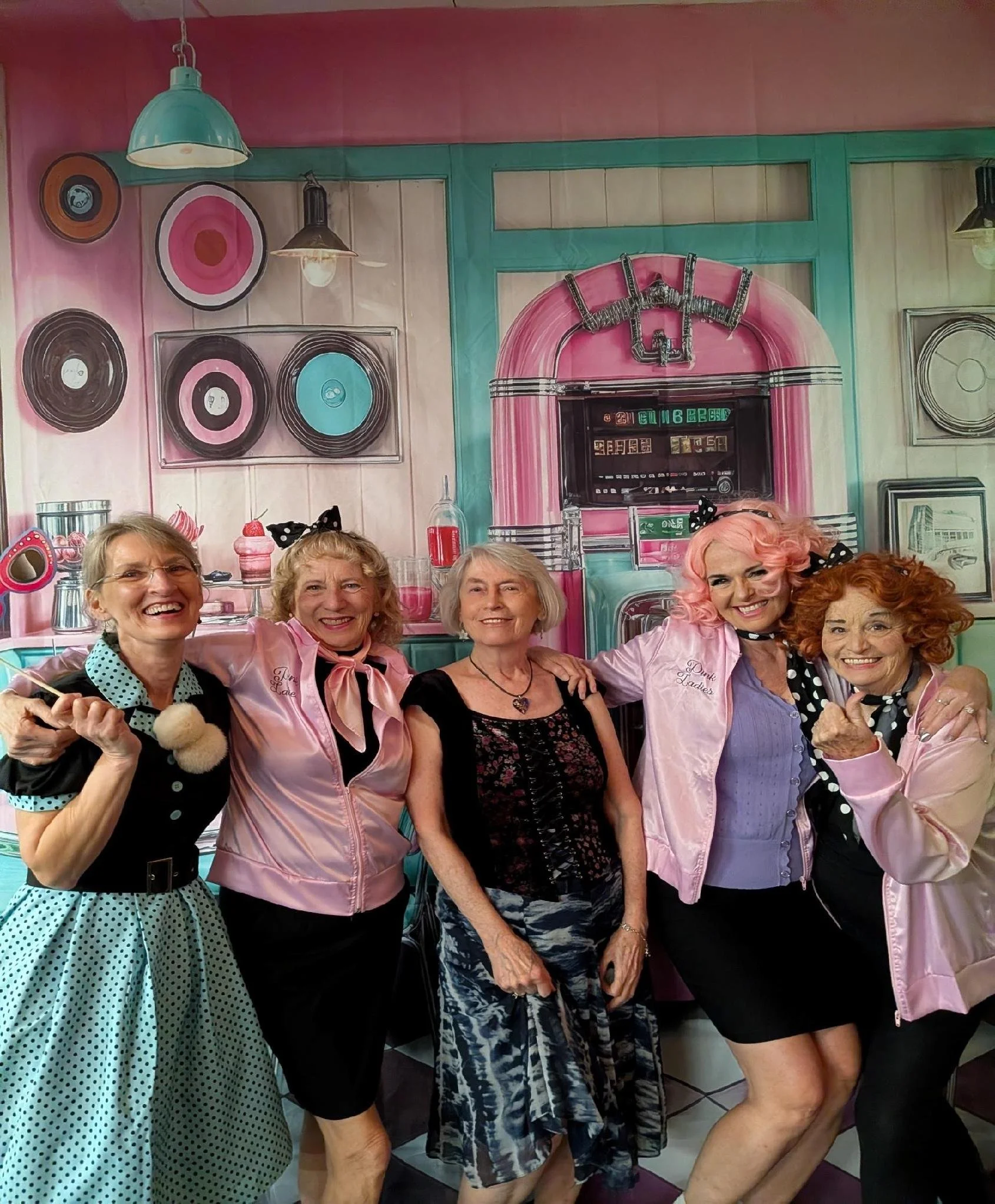 Group of women smiling and posing together in a decorated room with pink and teal accents, including art on the walls and vintage decor.