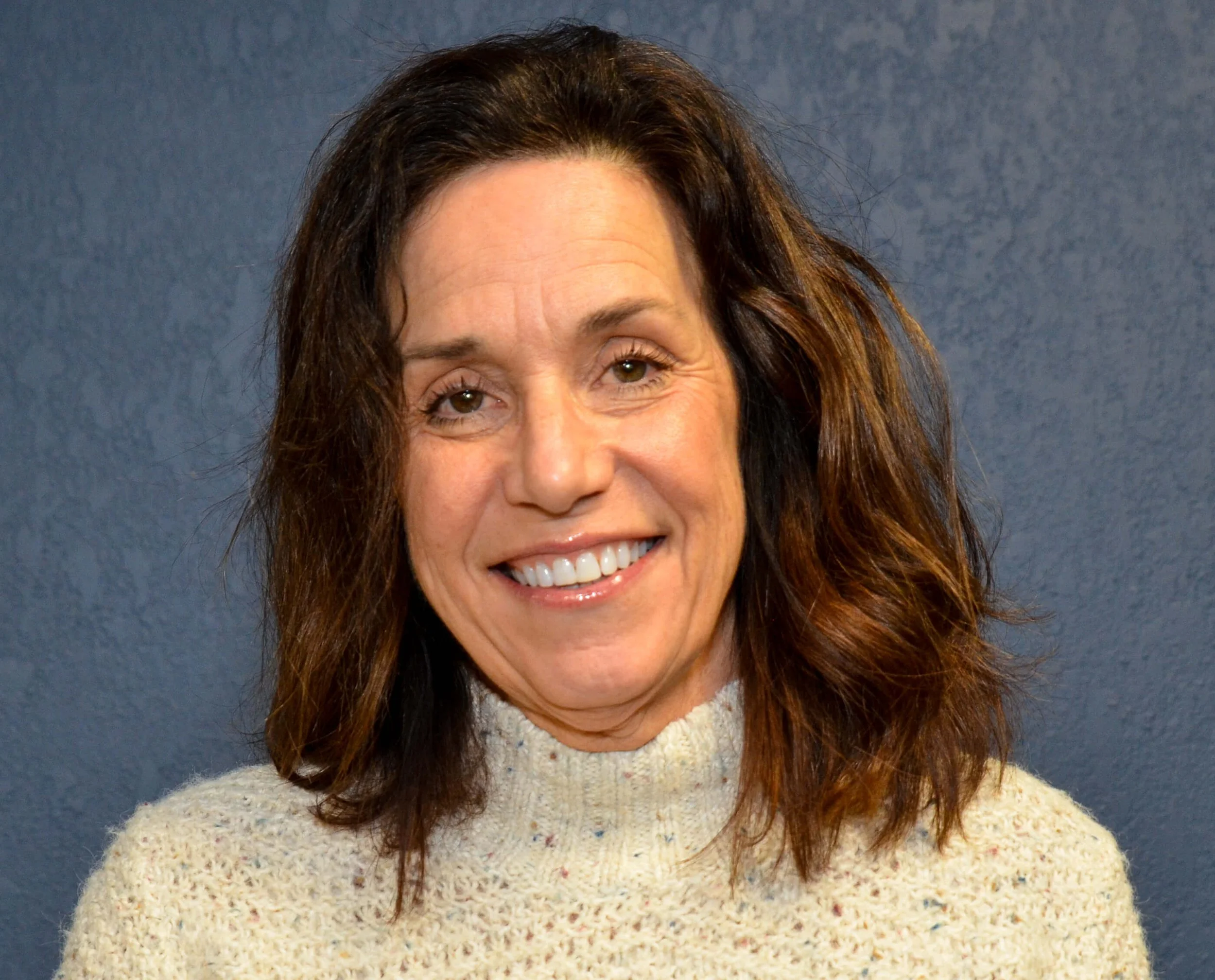 A smiling woman with brown, curly hair and light skin, wearing a cream-colored knit sweater, standing in front of a plain blue background.
