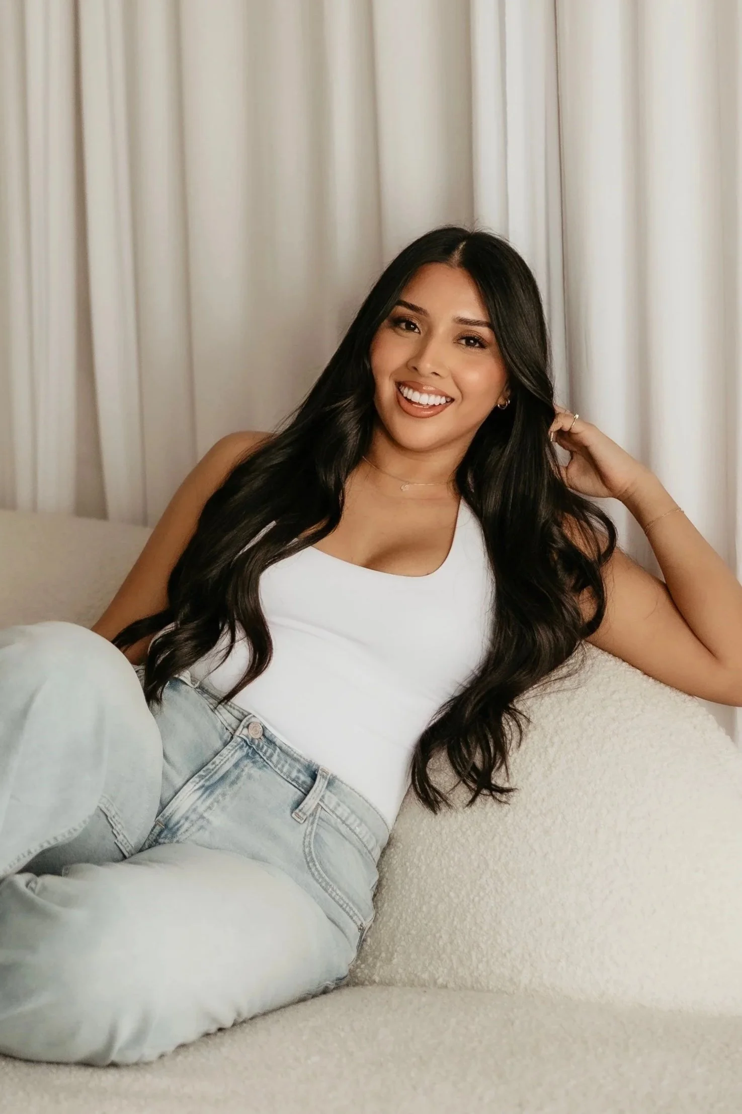 A smiling woman with long black hair sitting on a beige couch with a white cushion, in front of closed white curtains, wearing a white sleeveless top and light blue jeans.