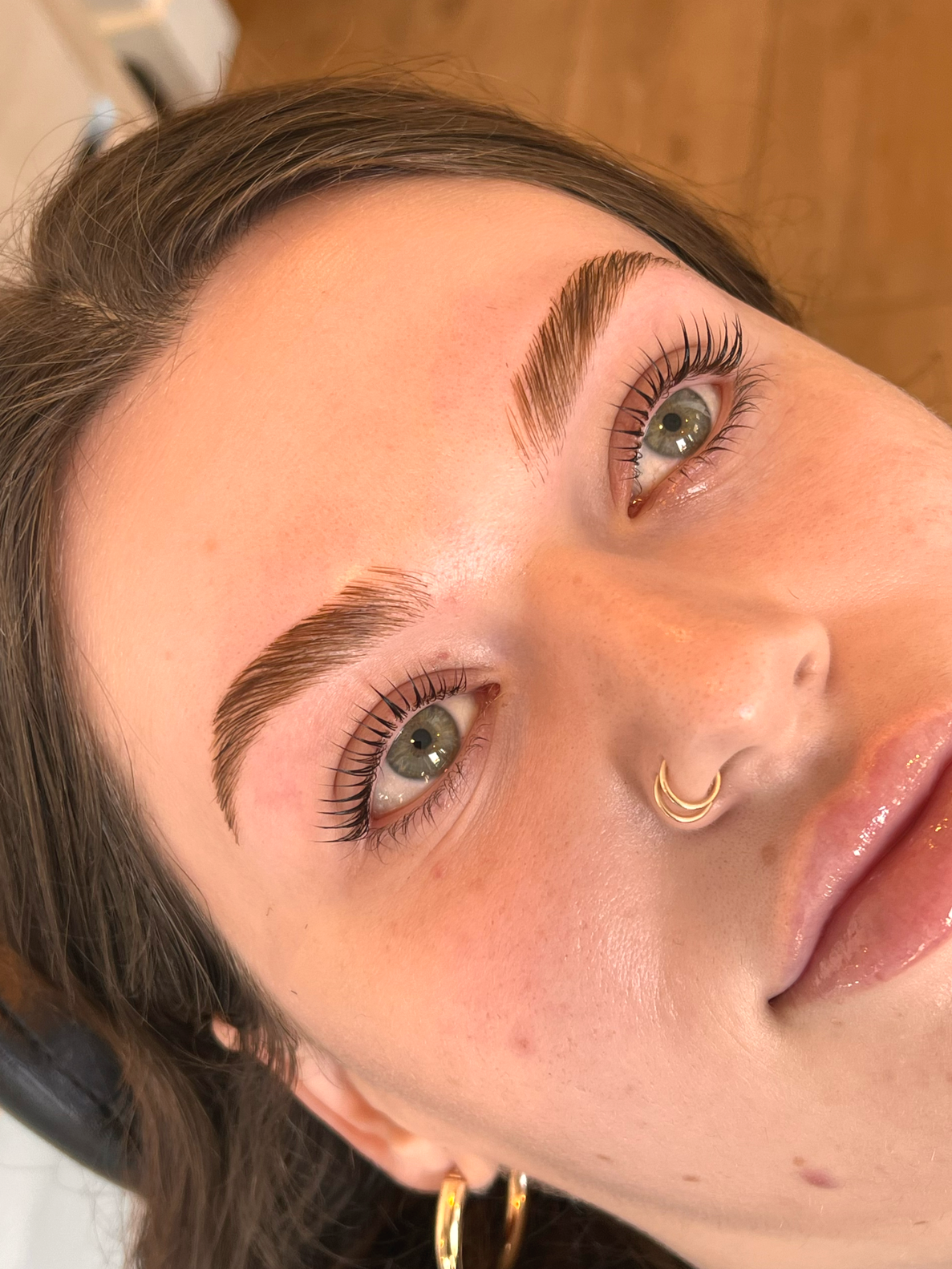 Close-up of a woman's face, focusing on her eyebrows, eyes, and nose. She has well-groomed eyebrows, green eyes, long eyelashes, and a gold septum piercing. Her skin is clear with a few small blemishes.