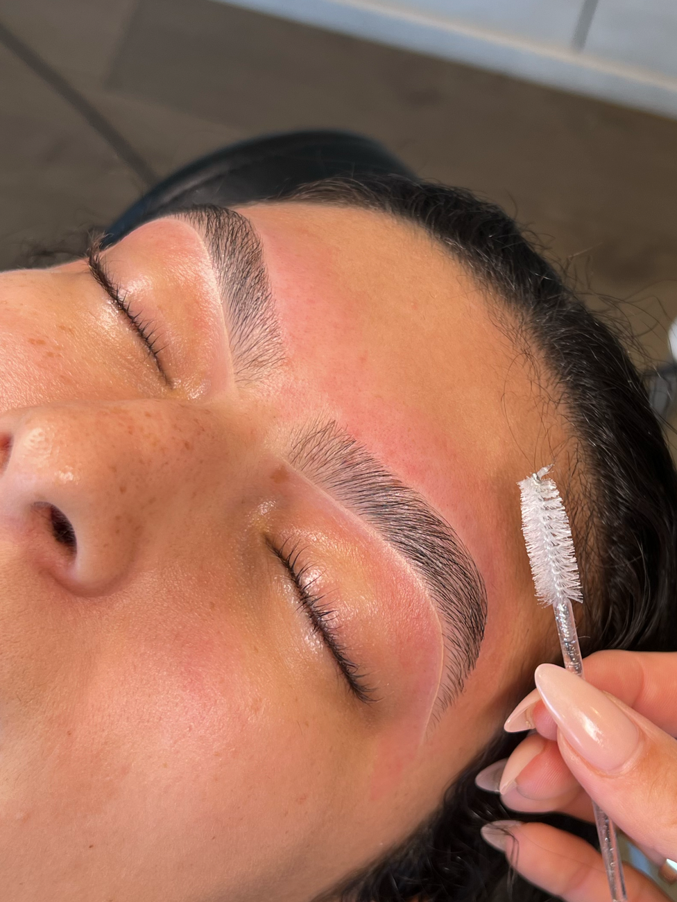Close-up of a woman lying down with her eyes closed as an aesthetician applies brow gel with a spoolie brush to her eyebrows.