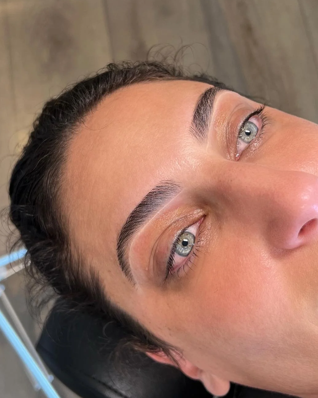 Close-up of a woman's face showing freshly done eyebrows, with her eyes open, lying on a black surface with a blurred background.