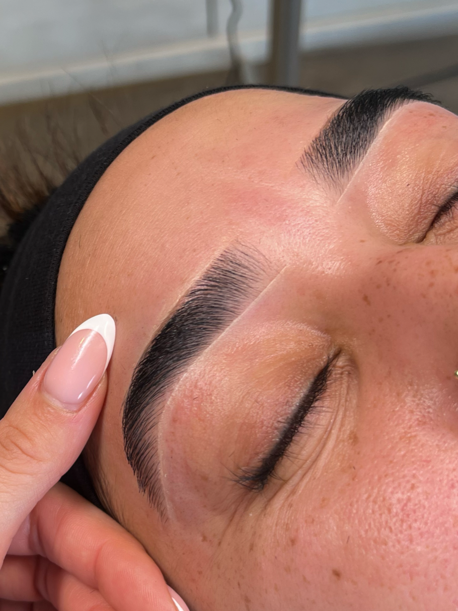 Close-up of a woman's face with freshly microbladed eyebrows, lying down with eyes closed, and a hand gently resting near her temple.