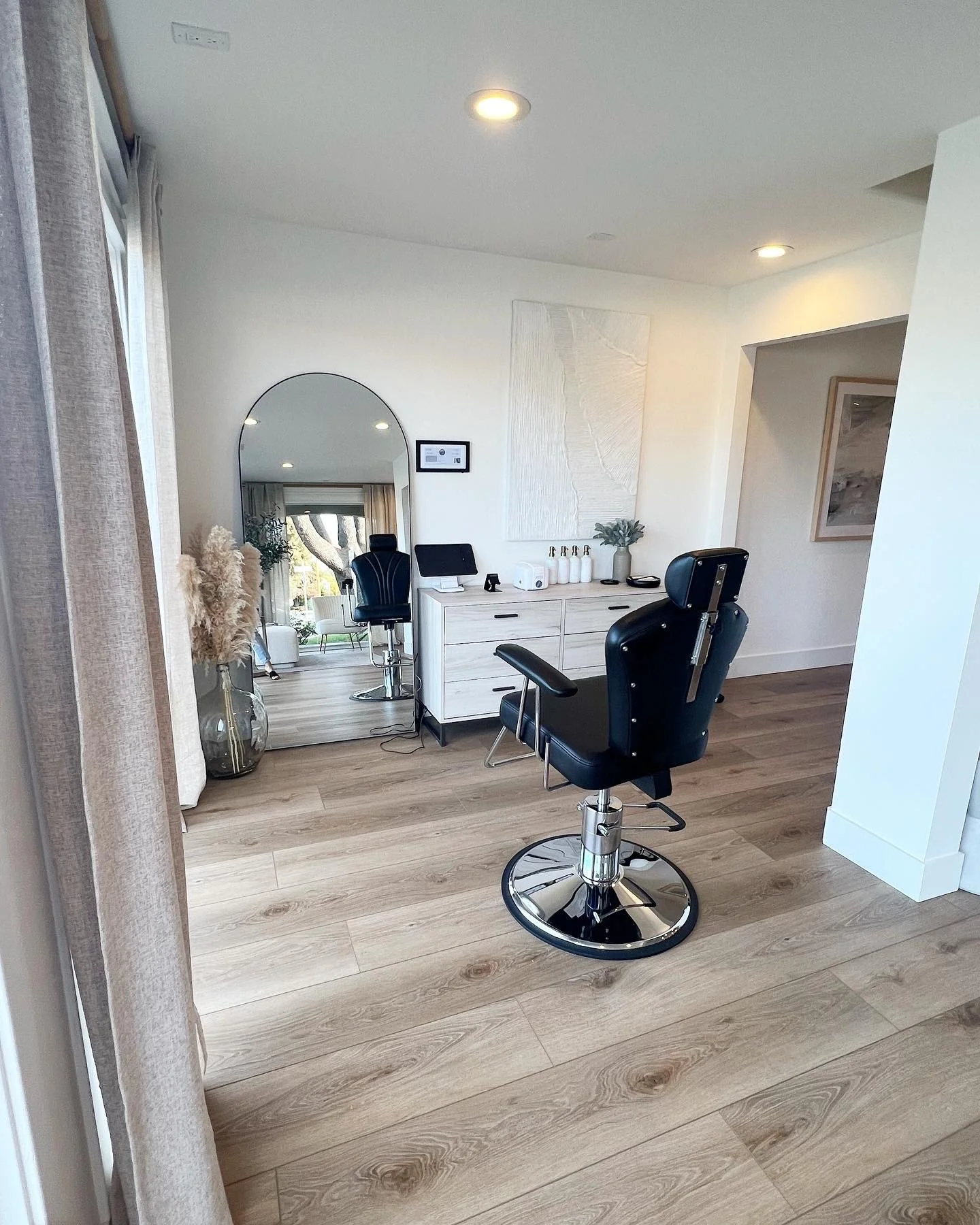 A modern hair salon with a black barber chair, a large mirror, and a white dresser with hair products, a plant, and a small monitor.