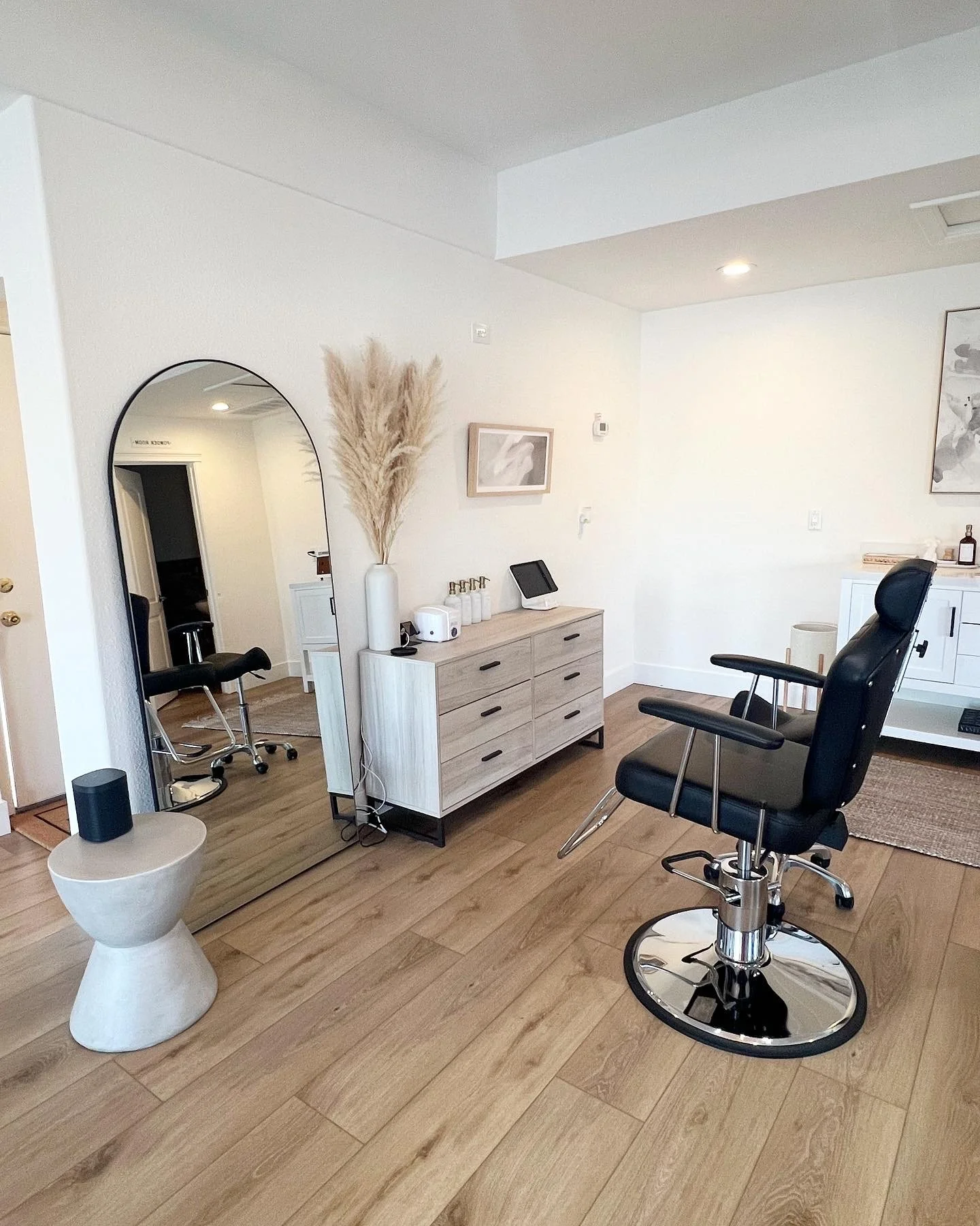 A modern living room with a large black recliner chair, a wooden dresser, a mirror, and a white cabinet, with framed artwork on the wall and decorative pampas grass in a white vase.