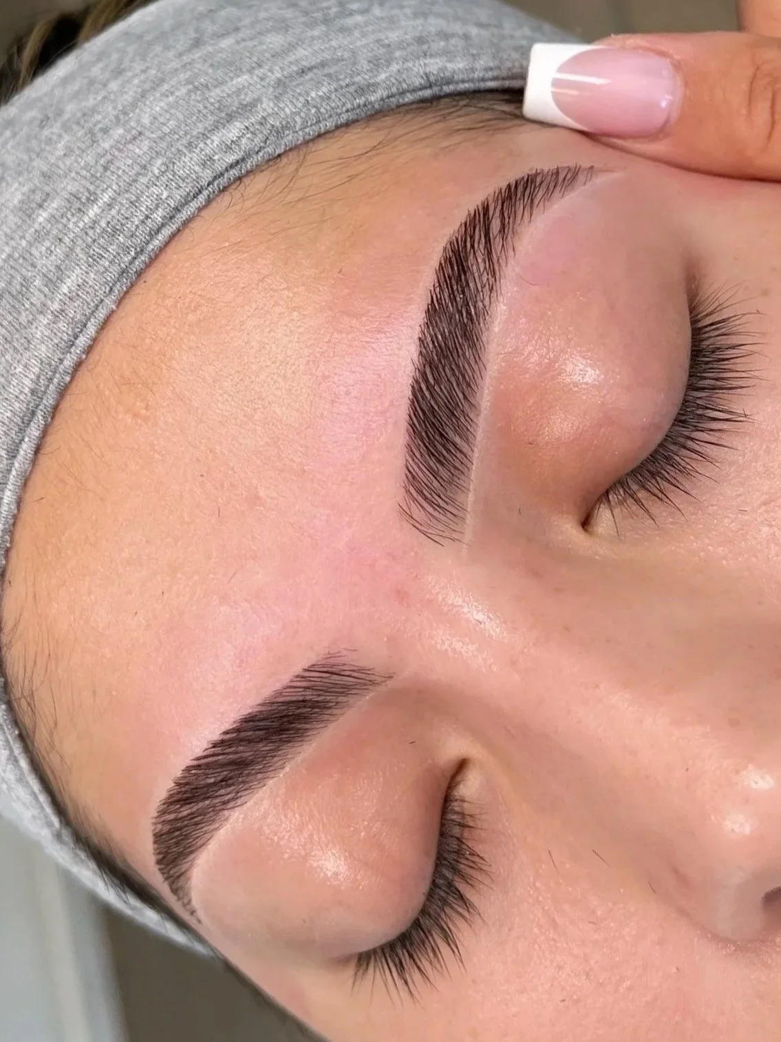Close-up of a person's face with eyes closed, showing freshly done eyebrows and long eyelashes, with a hand gently touching the forehead