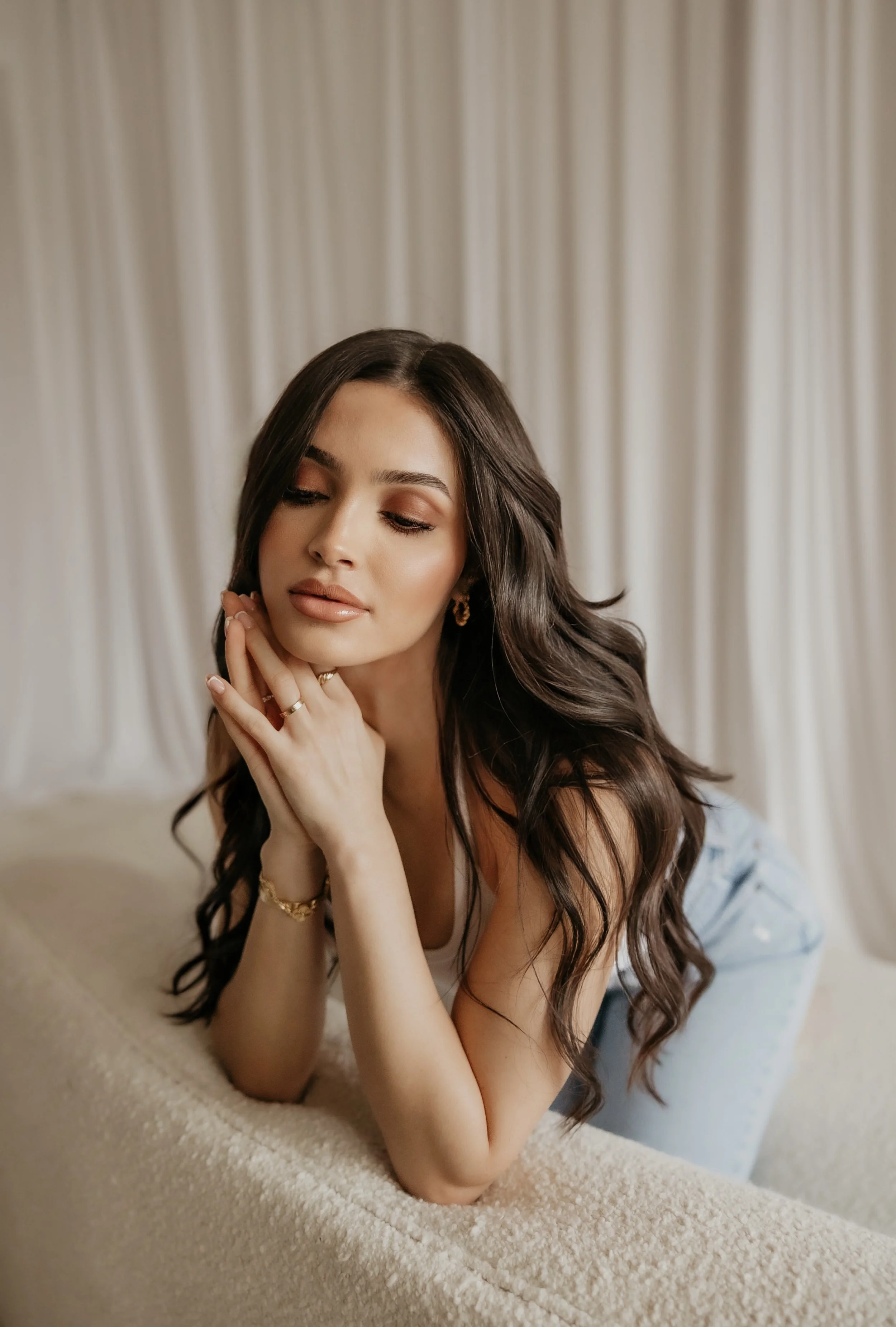 A young woman with long dark hair and makeup, resting her face on her hands, looking down with a gentle expression, in front of a beige curtain backdrop.