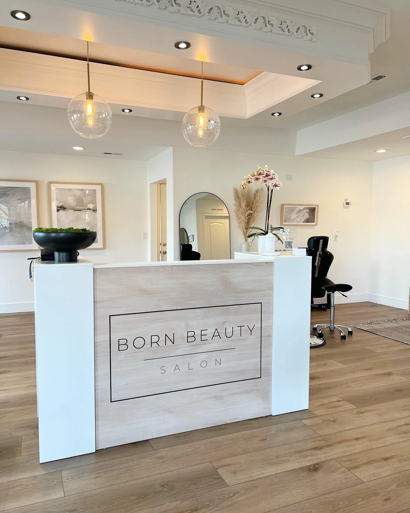 Reception desk at Born Beauty Salon with decorative items like a black bowl, white vases, and artwork on the walls, in a modern, well-lit interior with wooden flooring.