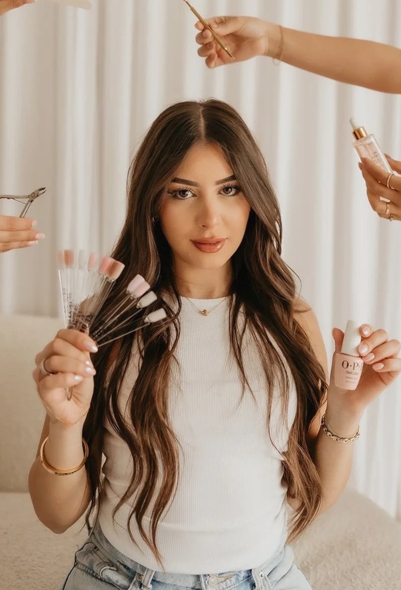 Woman with long wavy brown hair getting her nails done, holding a nail polish bottle, with multiple nail polish color samples in her other hand, while nail technicians work on her nails.