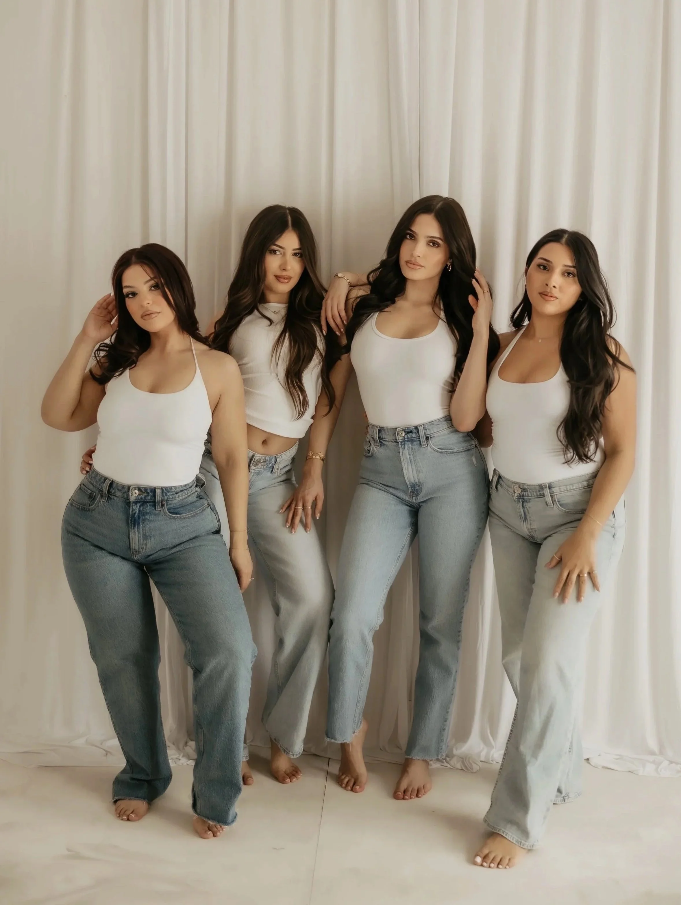 Four women standing in front of a white curtain, dressed in casual white tops and jeans, posing for a photo.