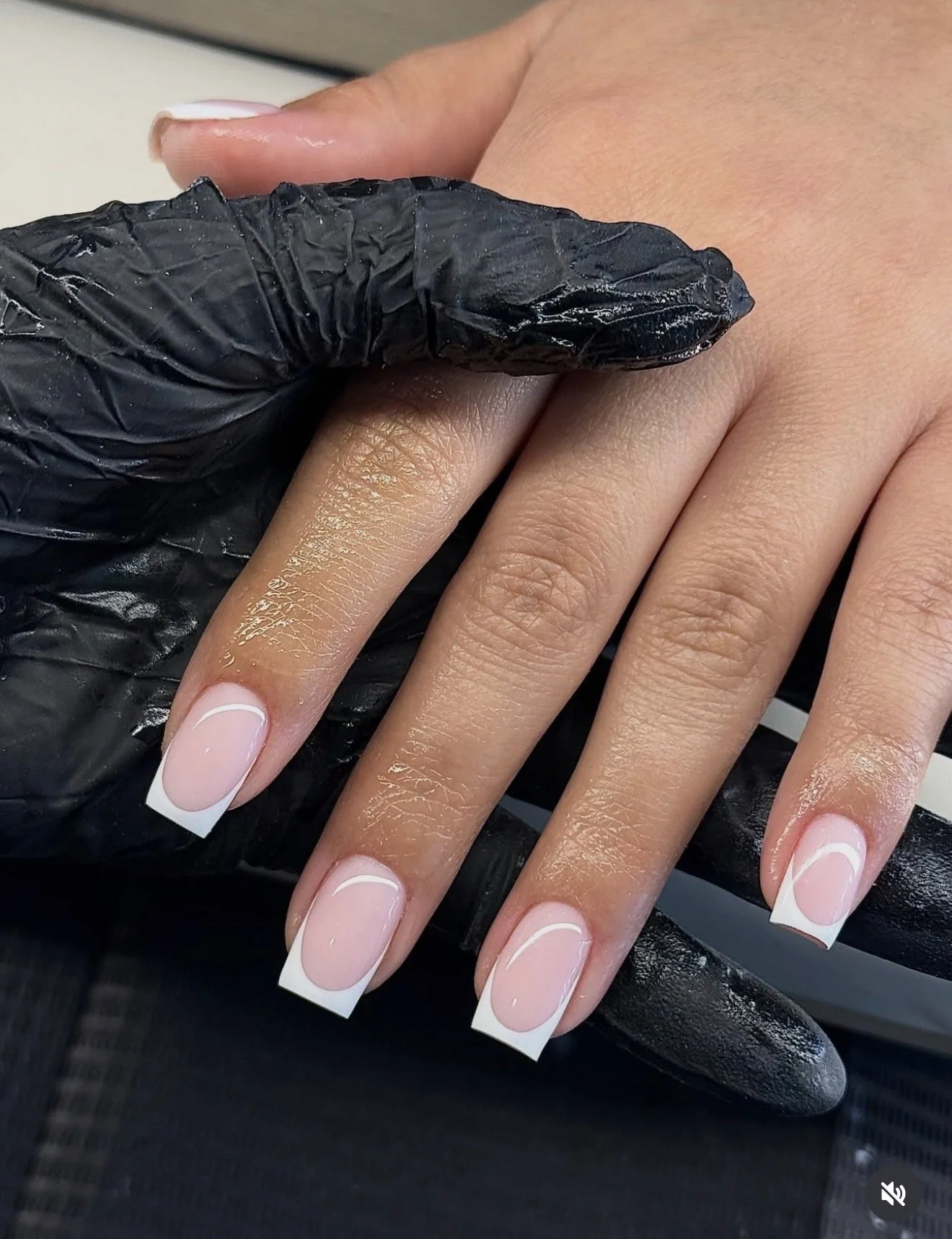 Close-up of a person's hand with freshly painted French manicure nails, held by a person wearing black gloves.
