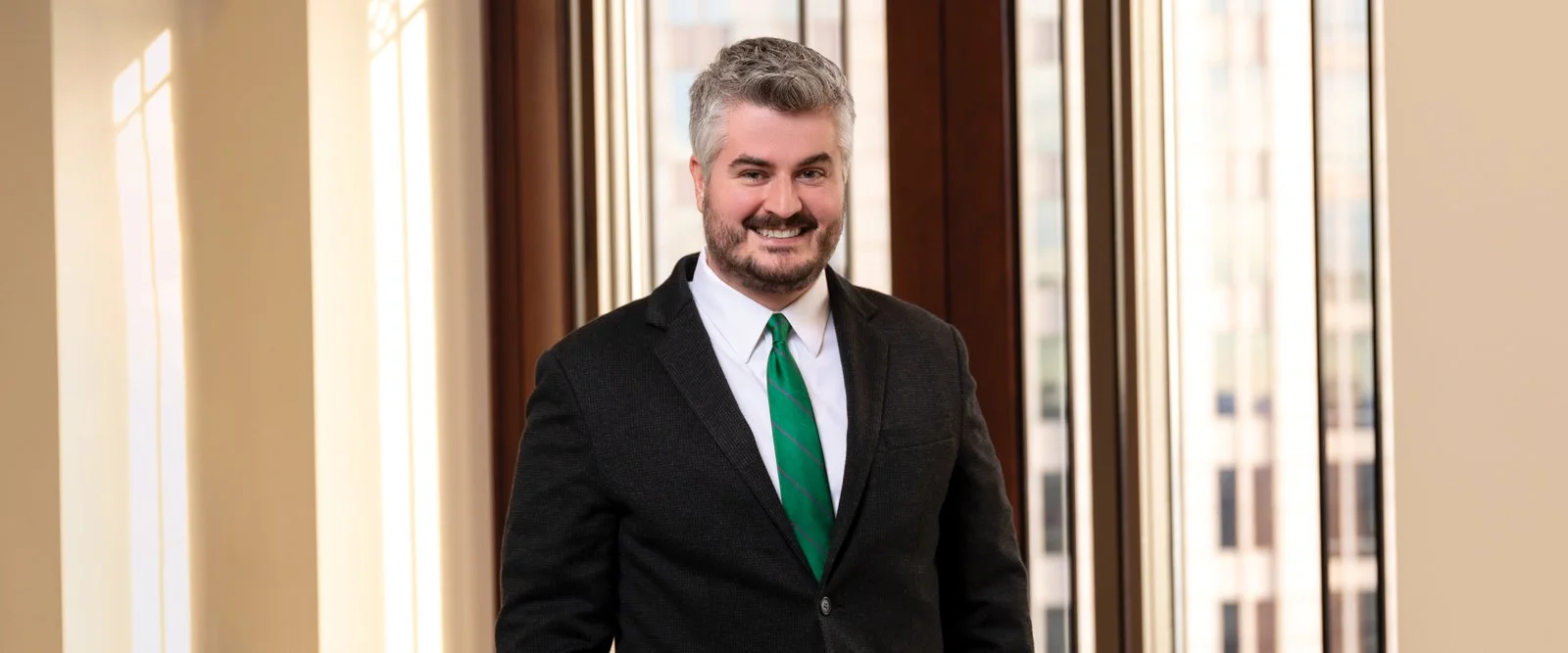 A smiling man with gray hair and a beard, wearing a black blazer, white shirt, and green tie, standing in front of large windows in an office setting.