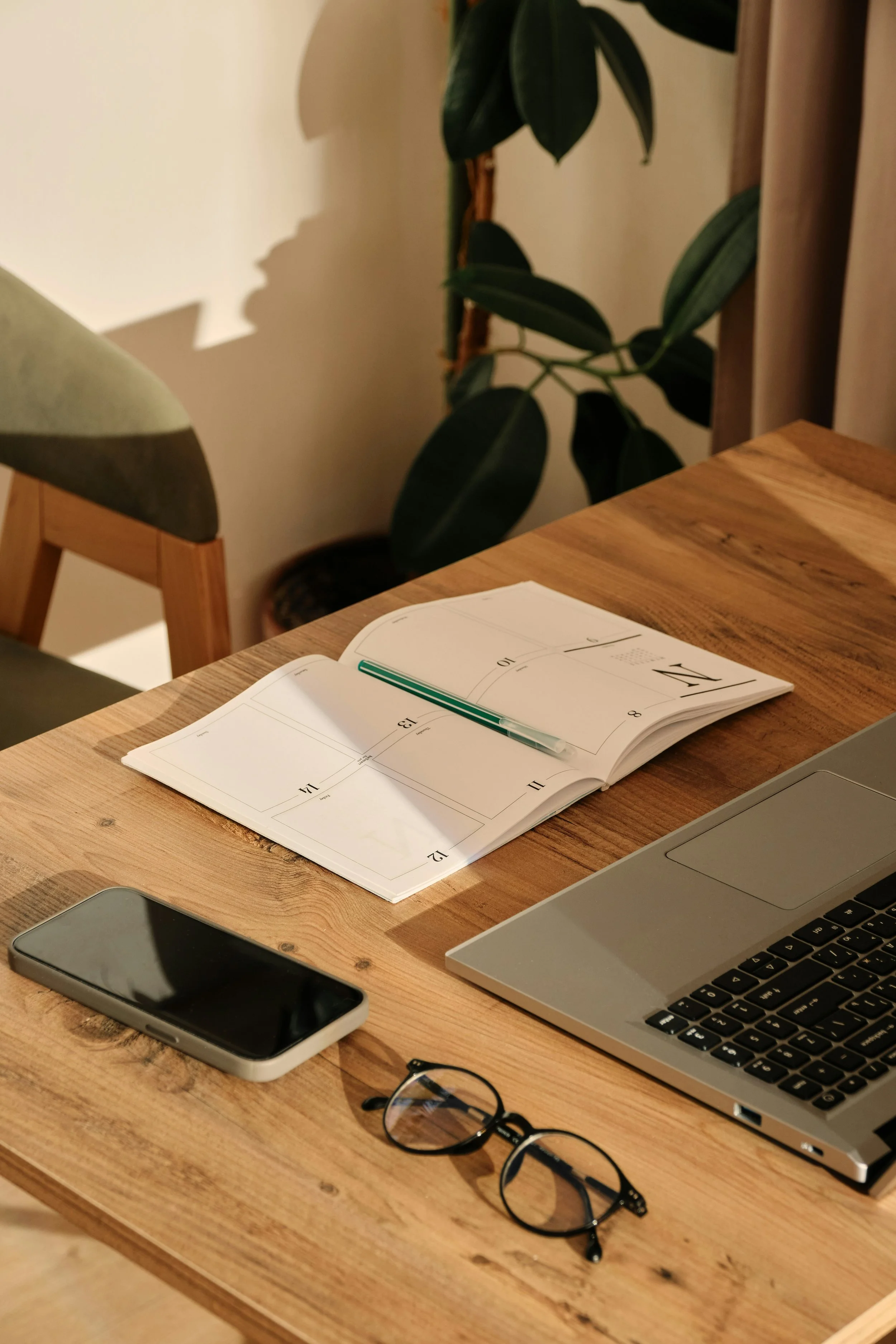 A wooden desk with an open weekly planner, a green pen, a smartphone, reading glasses, and a laptop, suggesting an organized and intentional approach to work.