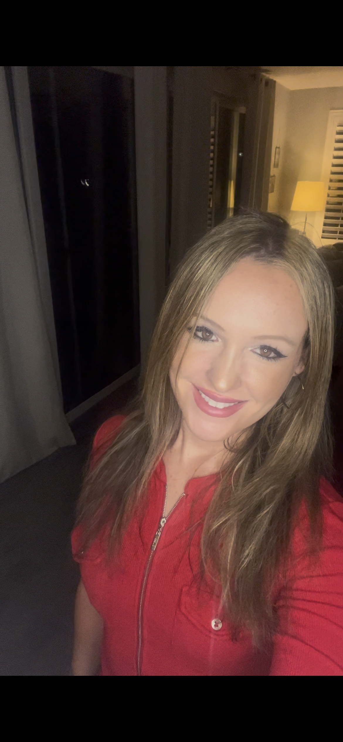 Portrait of a smiling woman with long brown hair, wearing a red zip-up jacket, inside a room with warm lighting and wooden furniture.