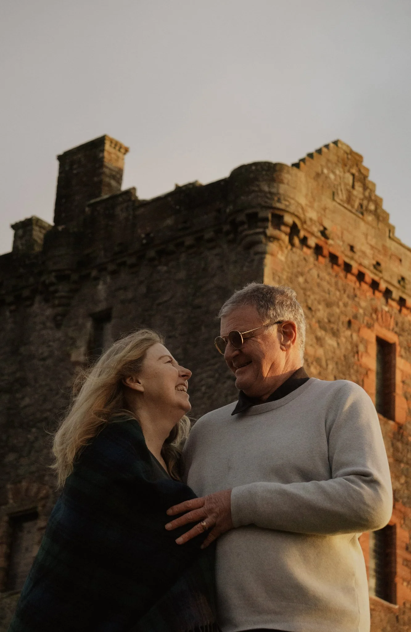 A smiling couple embracing happily in front of the historic stone ruins of a Scottish castle tower house.
