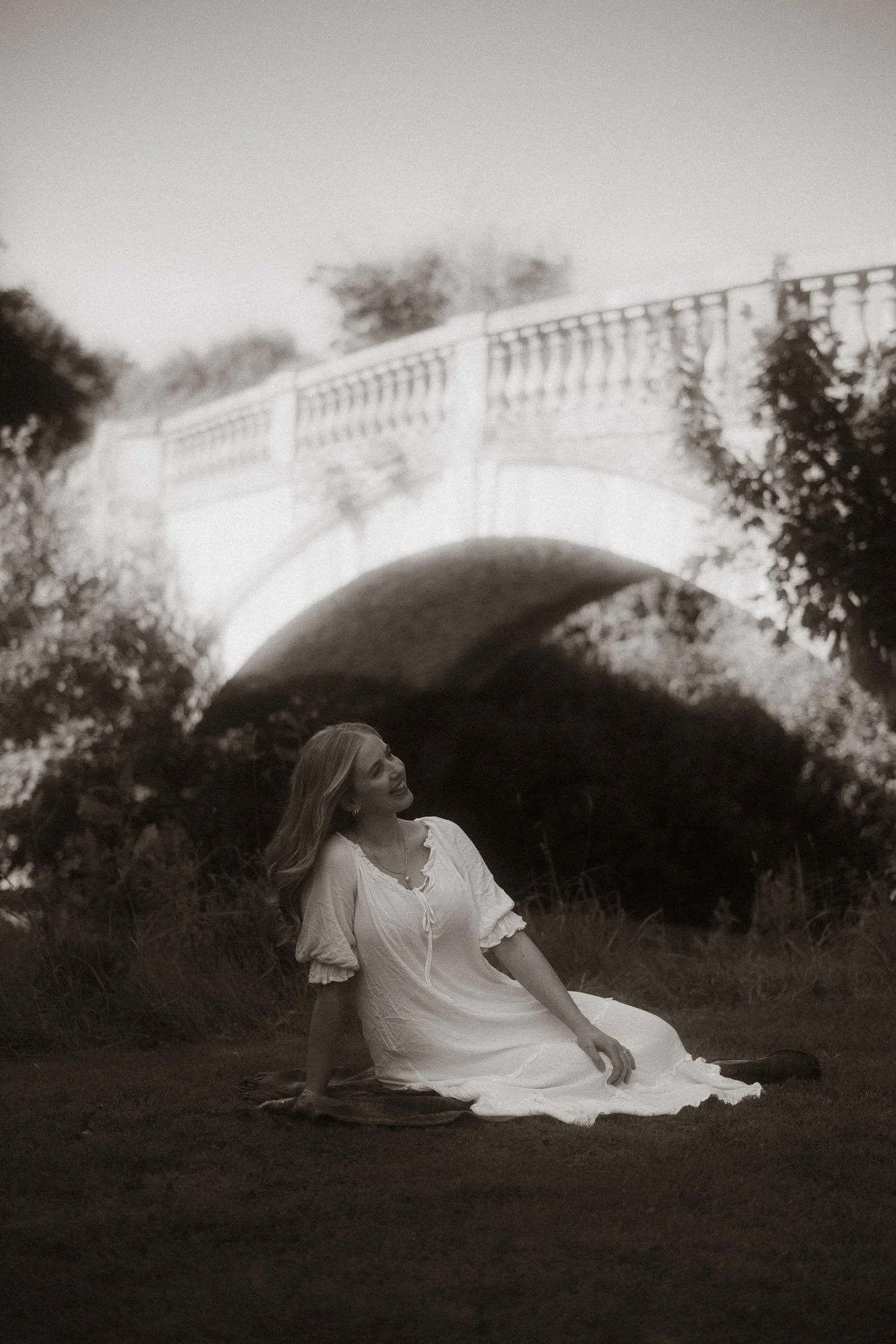 A vintage-inspired black and white fine art portrait of a woman in a dress sitting beneath a large historic stone arch bridge in Scotland.