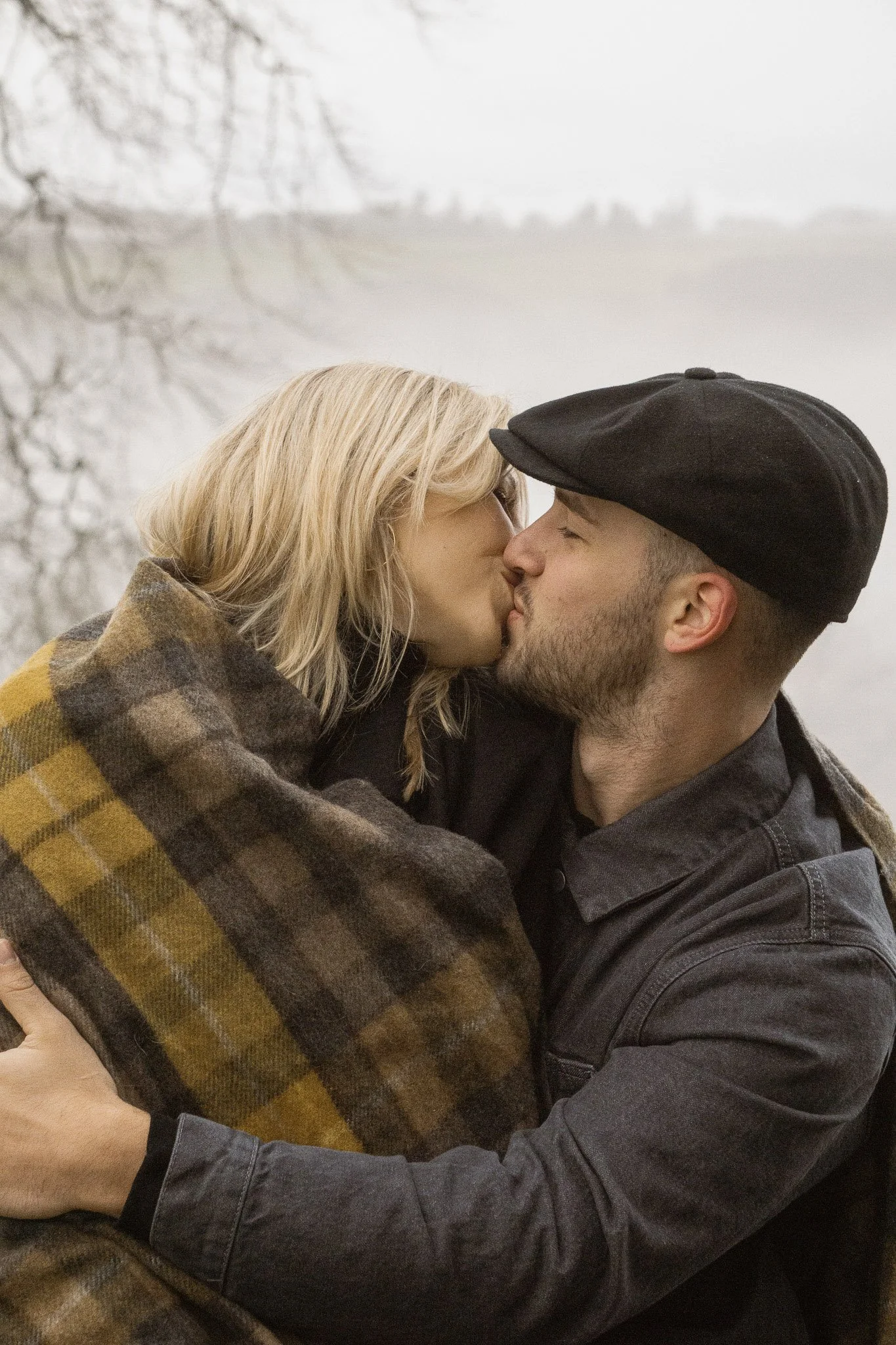 A close-up romantic portrait of a couple kissing while wrapped in a plaid tartan blanket with a misty loch in the background.