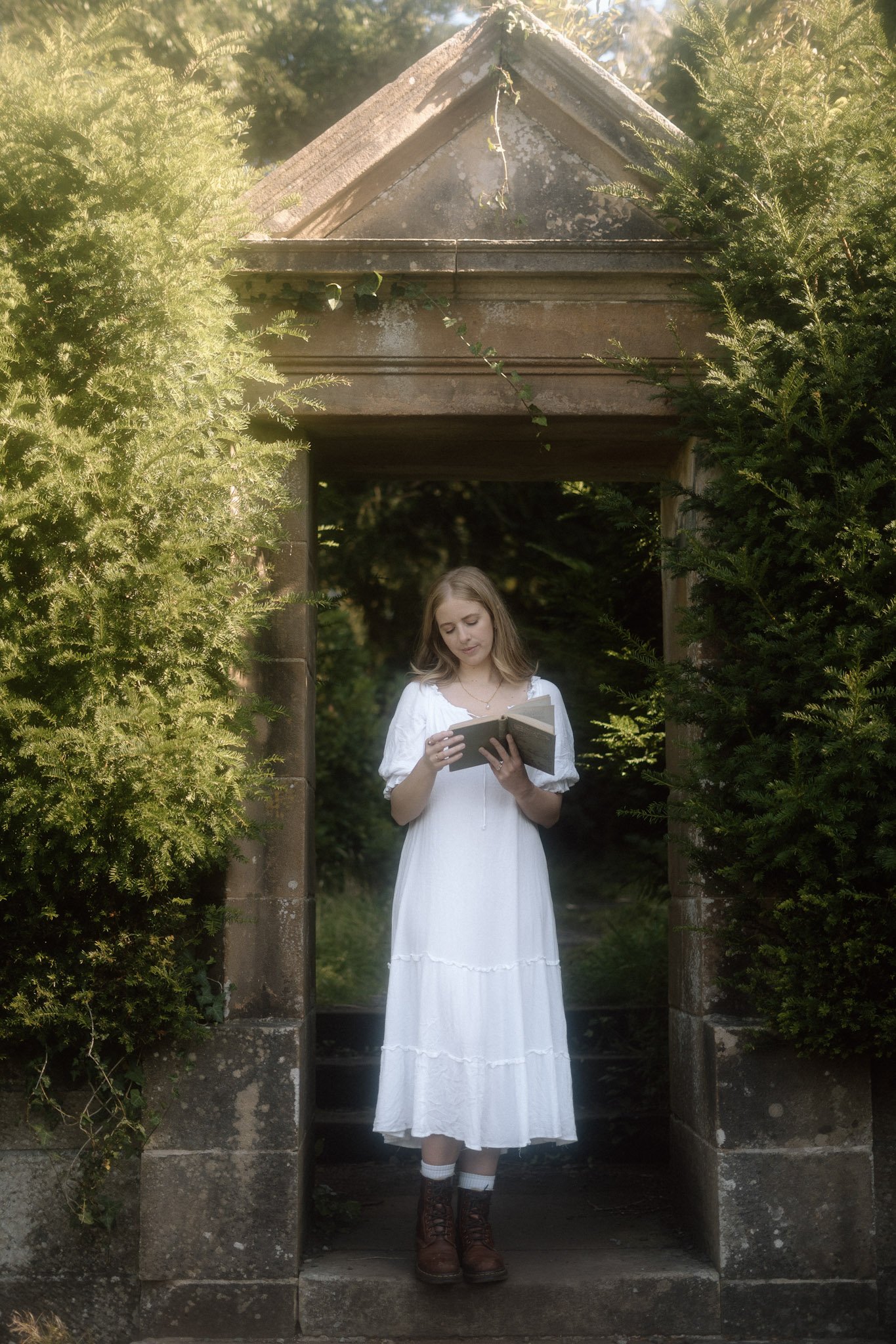 A woman in a flowing white dress standing framed inside an old, historic stone archway in Scotland surrounded by ivy and ivy-covered garden walls.