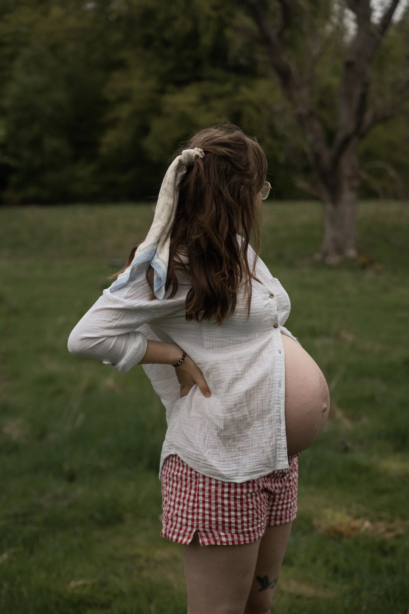 A pregnant woman wearing a white dress looking out thoughtfully across a vast, green grassy field during a Scottish Highlands maternity session.