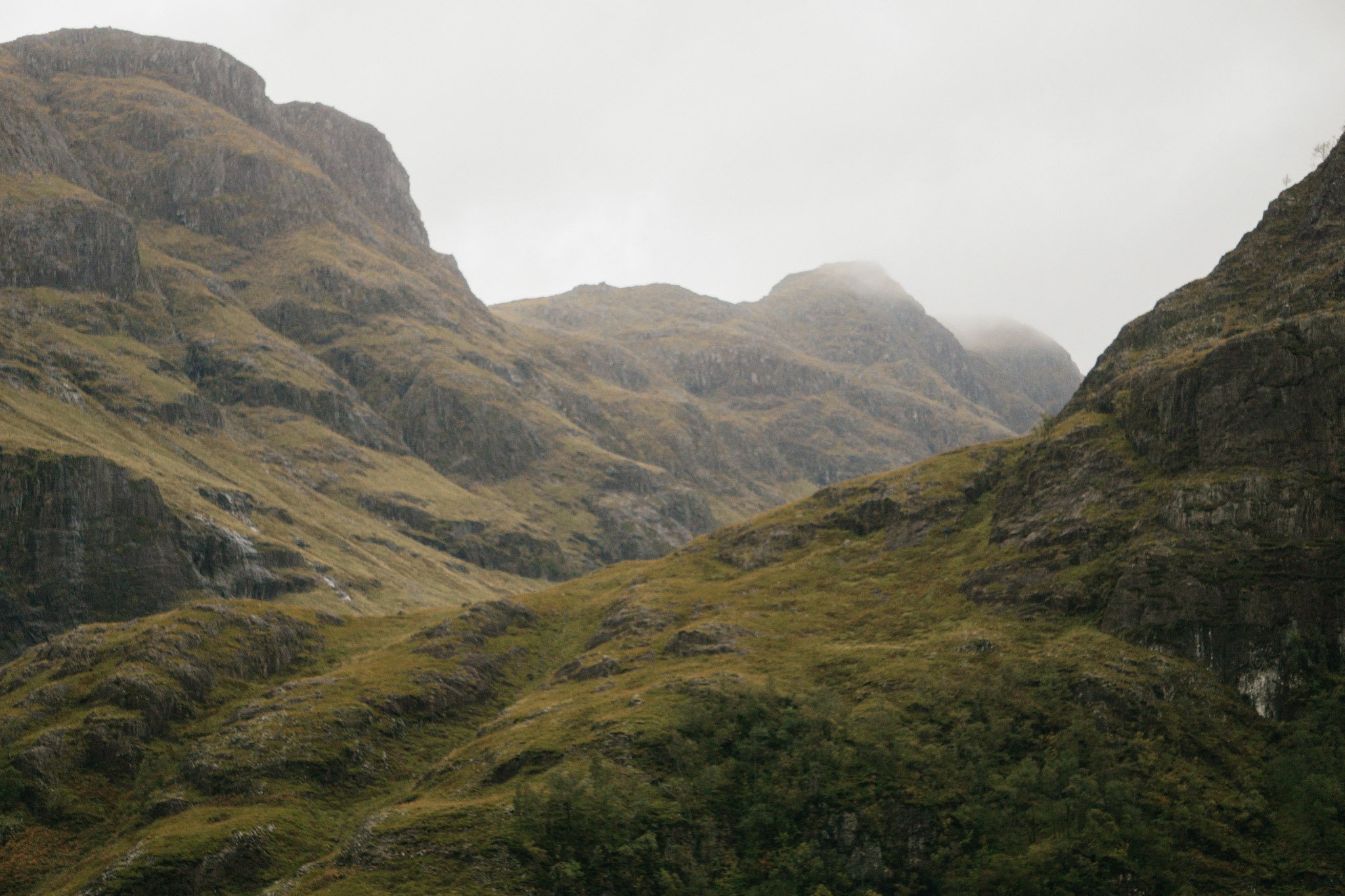 A sweeping, cinematic landscape view of the dramatic, rocky peaks of Glencoe under an atmospheric overcast sky.