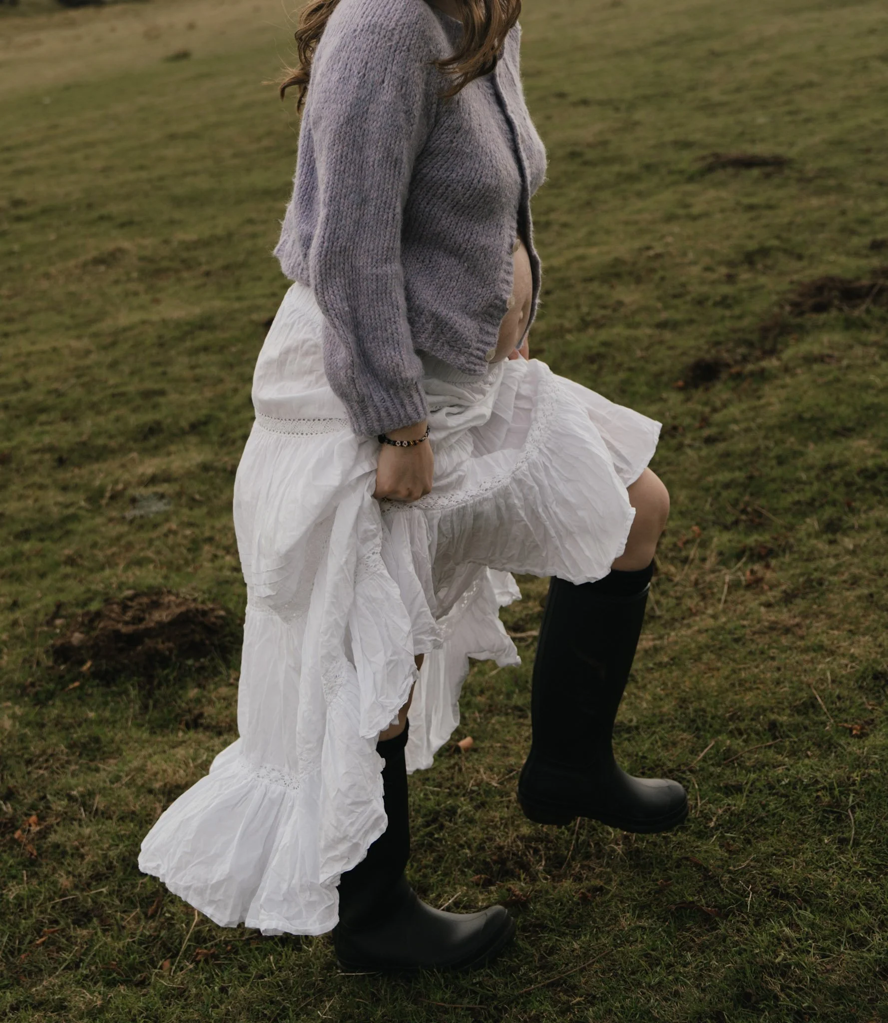 A woman wearing a long white bohemian-style skirt and black boots walking across a rugged grassy highland pasture.