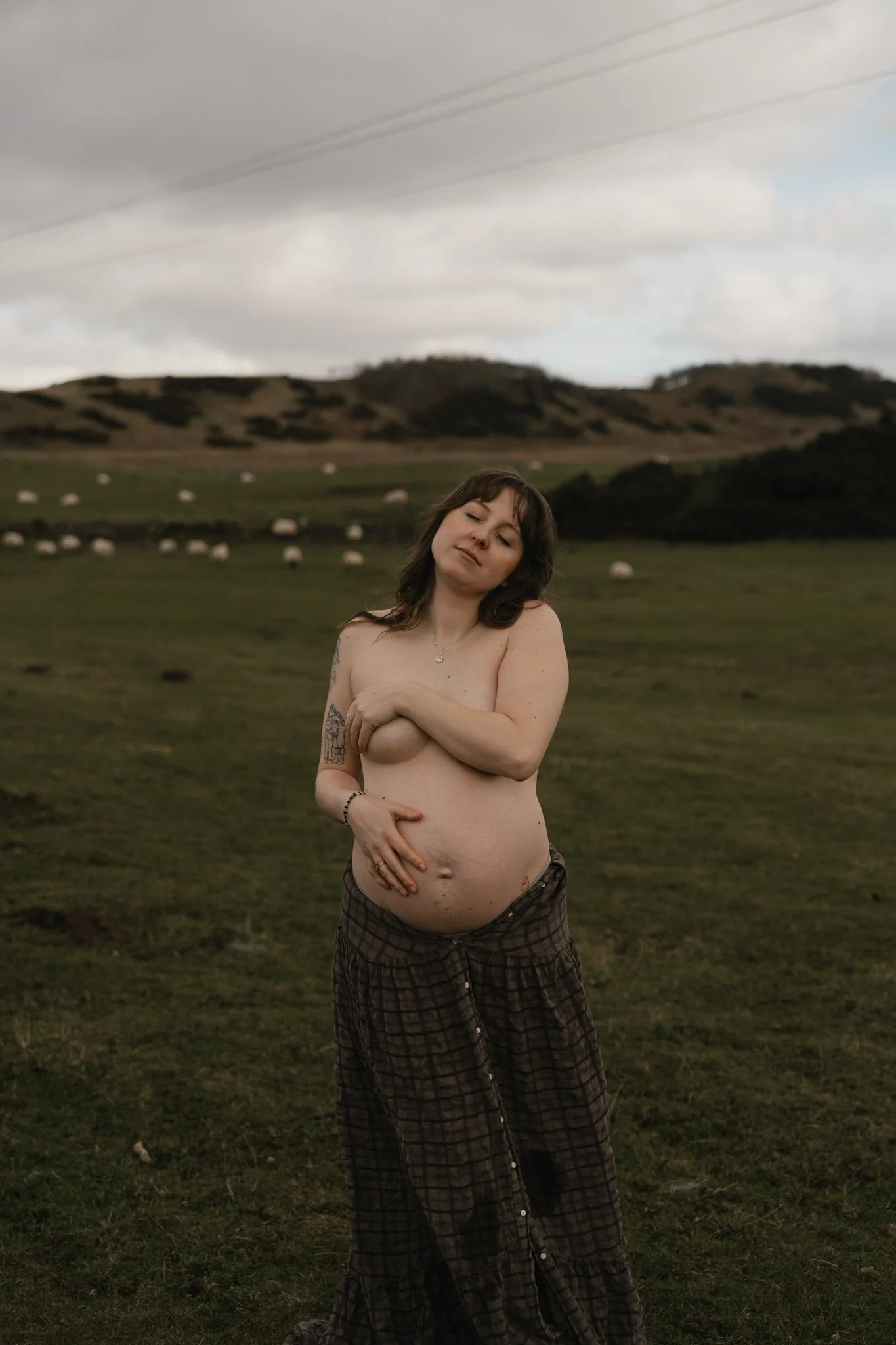 A beautiful maternity portrait of a woman standing in a rolling green Scottish field with sheep grazing quietly in the background.