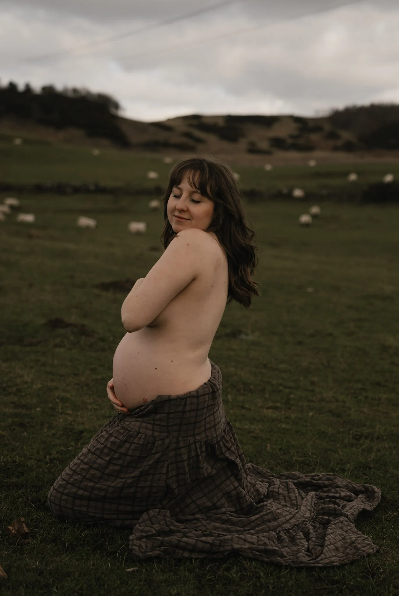 An artistic fine-art maternity portrait of a pregnant woman kneeling gracefully in a green Scottish field, with sheep grazing on the rolling hills in the background.