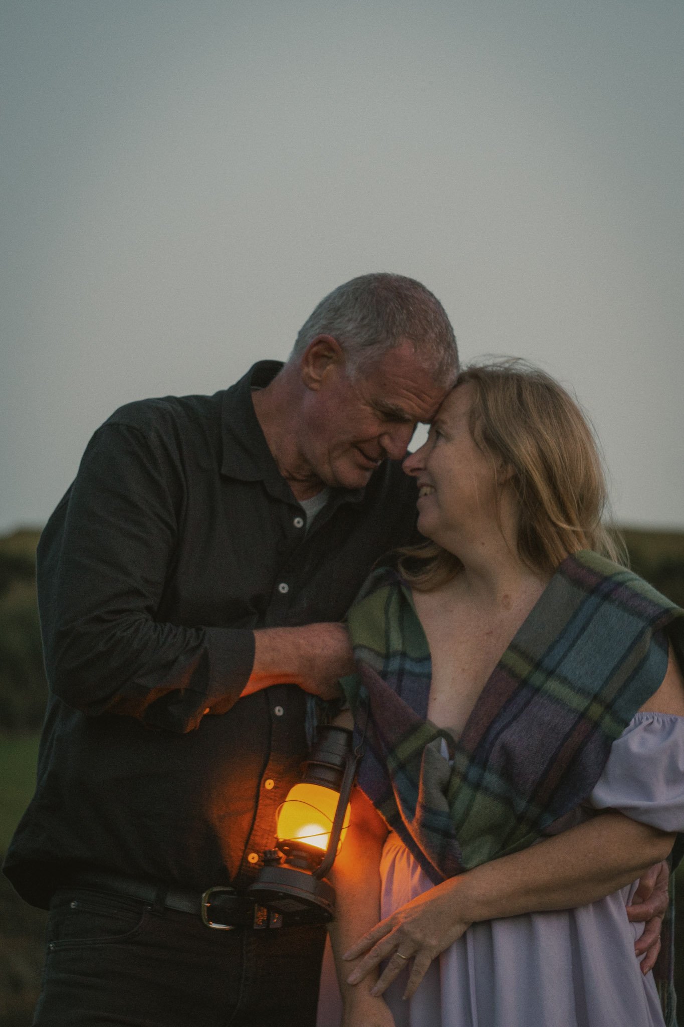 A warm, intimate close-up of a couple standing close together at dusk, illuminated entirely by the amber glow of a handheld lantern.