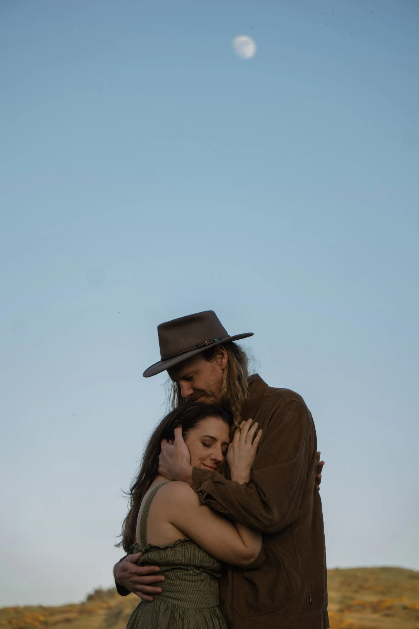 A man in a wide-brimmed hat holding a woman close under a pale blue twilight sky with a crescent moon visible.